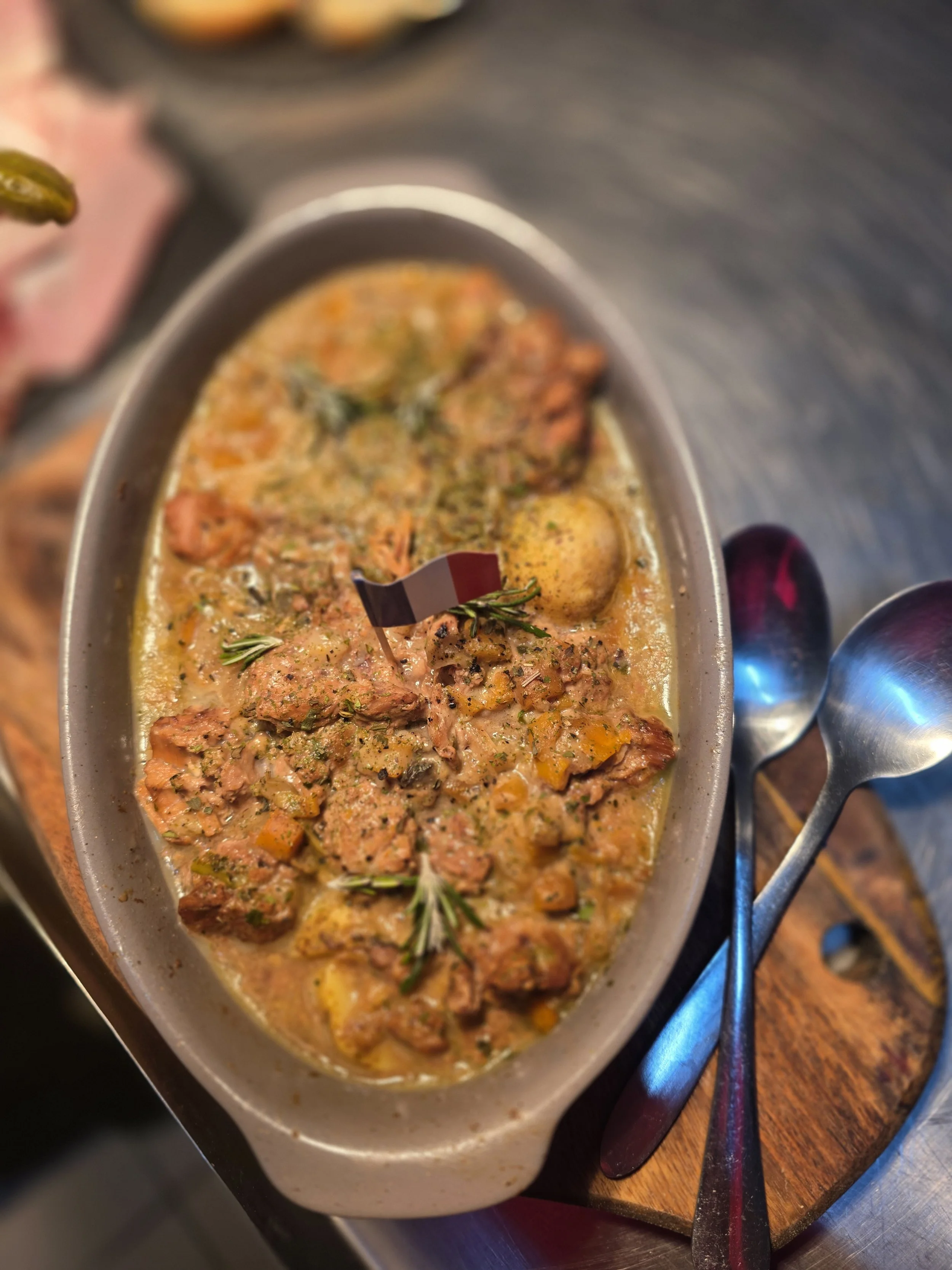 A oval ceramic dish filled with beef stew, garnished with sprigs of rosemary and a small French flag, served with two metal spoons on a wooden tray.