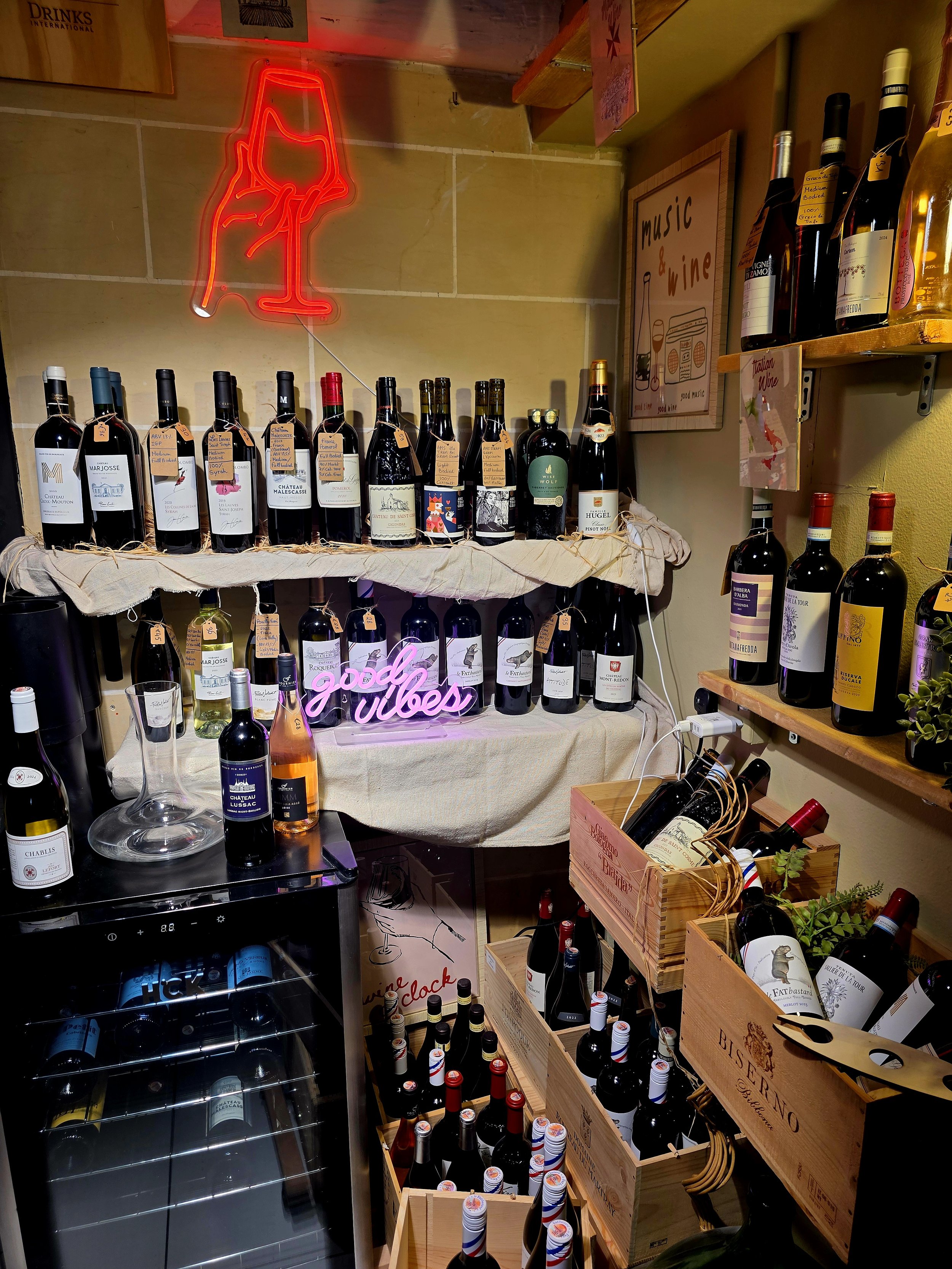 A display of wine bottles on shelves and in boxes in a cozy wine shop or bar. There is a neon sign shaped like a wine glass hanging on the wall, with a small pink sign that says 'good vibes' in front. A sign on the wall reads 'music & wine.' A wine r
