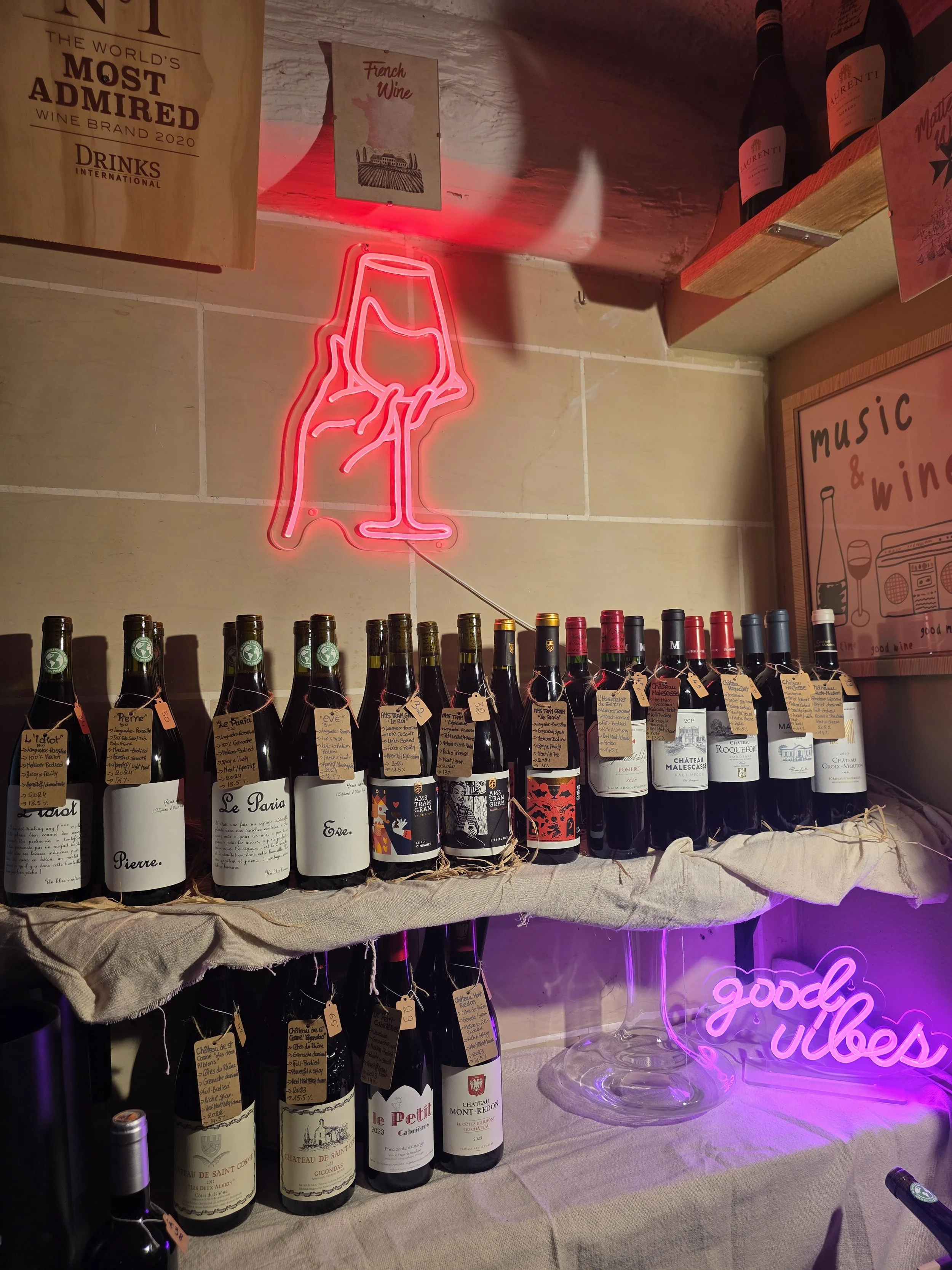 Display of various bottles of wine on a table with a neon sign of a wine glass and the words 'good vibes' in pink light, and other wine-related signs on the wall.