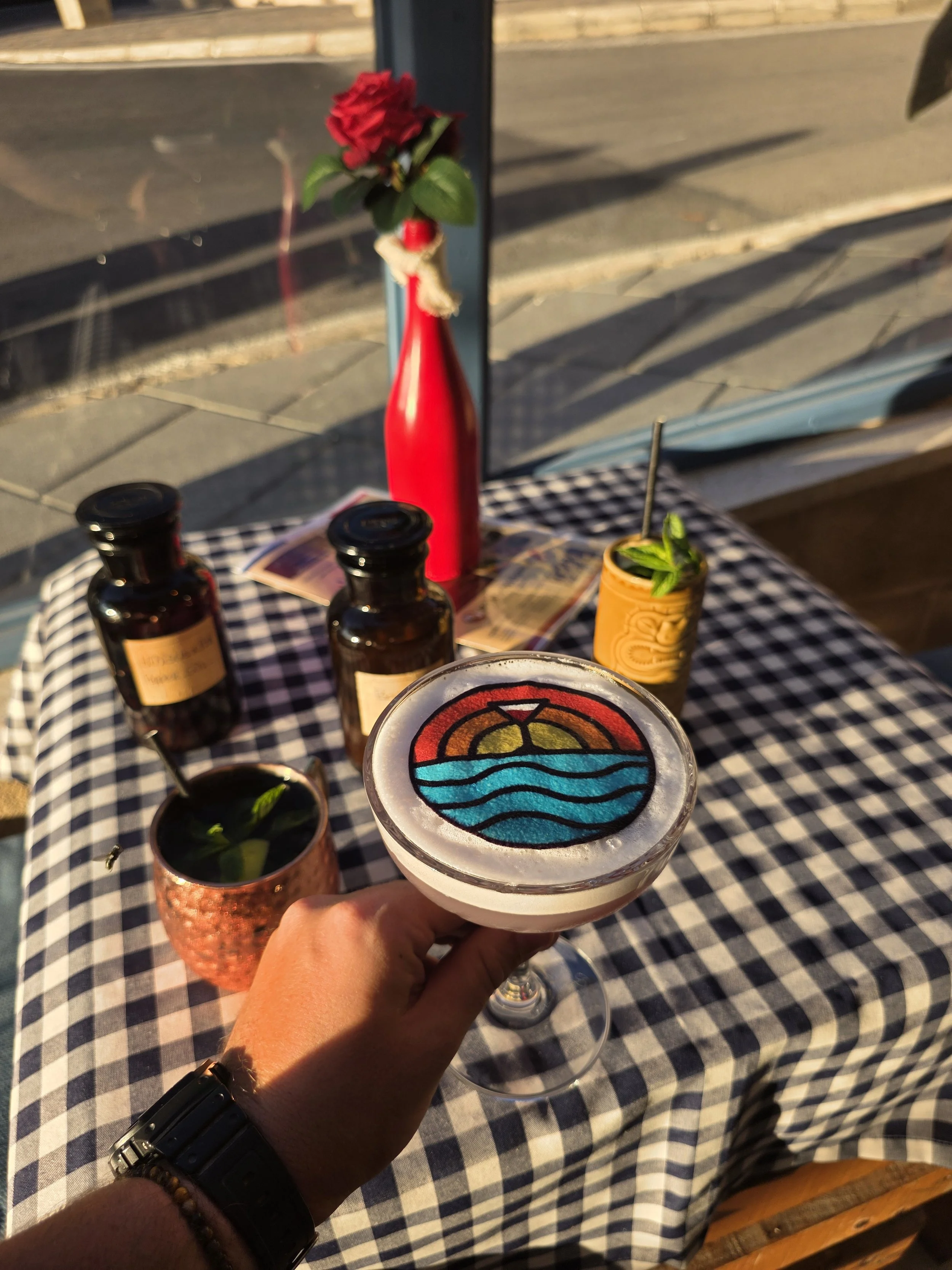 A person holding a martini glass with a colorful, stained glass-style sunset and ocean design on top. The table has a checkered tablecloth and is set with bottles, a copper cup with a drink, a red vase with a red flower, a yellow tumbler with a straw