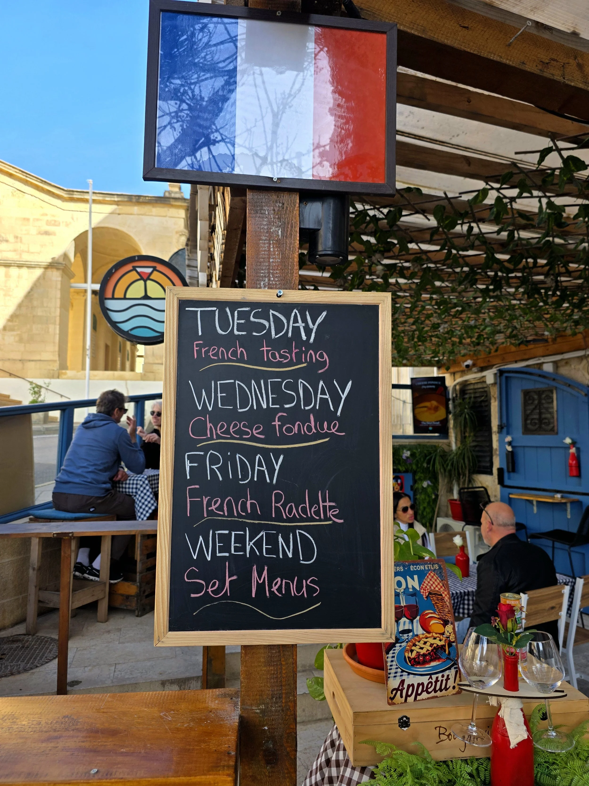 A chalkboard menu outside a restaurant listing special dishes for each day of the week, including French tasting on Tuesday, cheese fondue on Wednesday, and French Radlette on Friday, with set menus available on the weekend. There are also people din