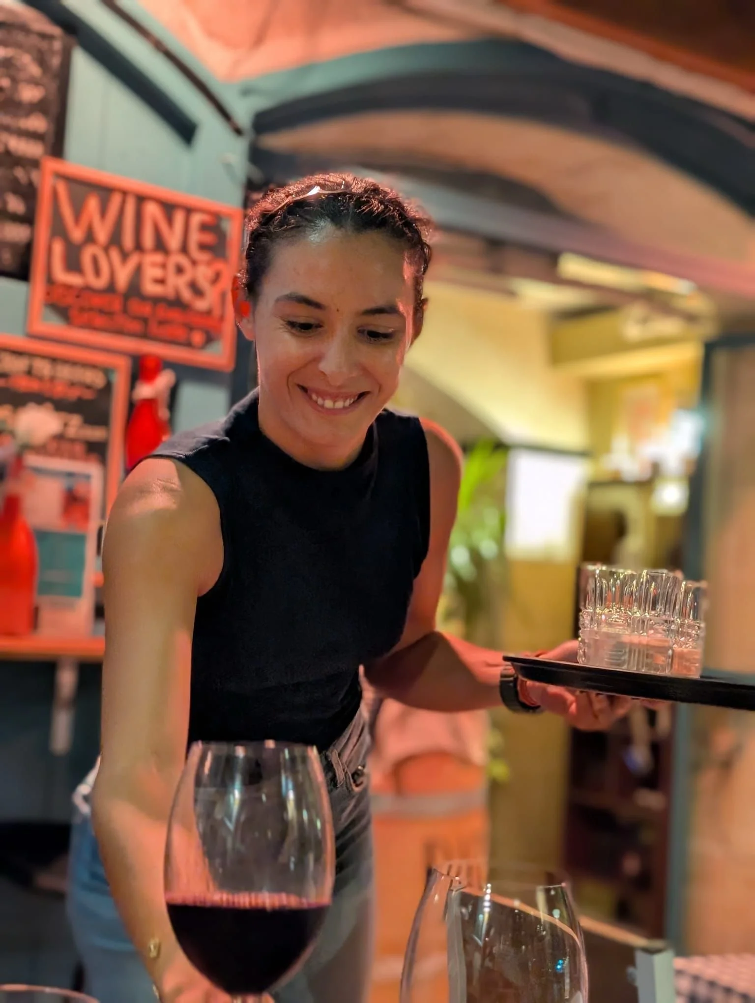 A woman serving a glass of red wine at a restaurant or bar, with a 'Wine Lovers' sign in the background.