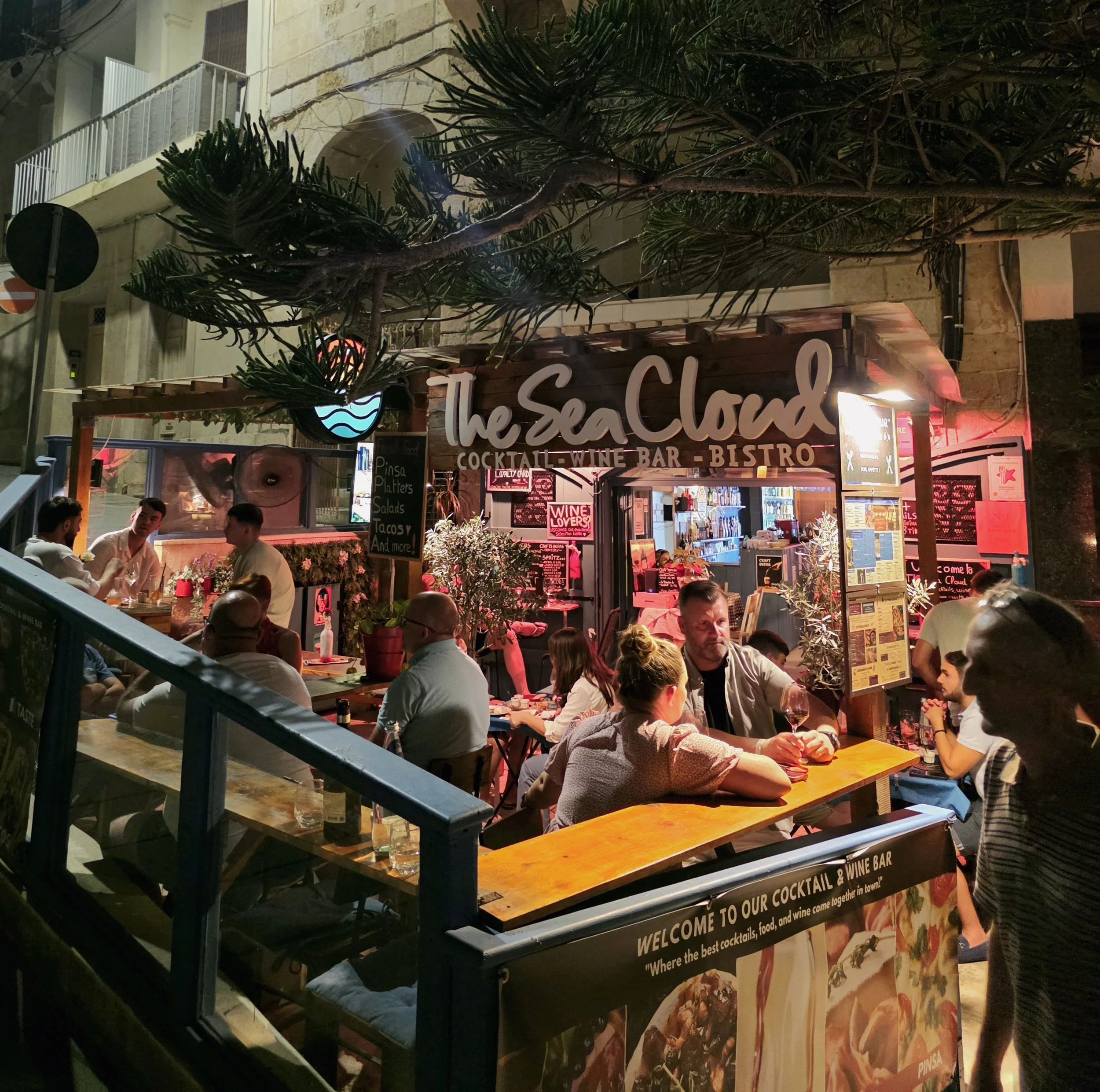 Night scene at a lively outdoor bar called 'The Sea Cloud,' with customers sitting at tables, some drinking wine, under a canopy of trees and string lights. The bar features a signboard and menu, with colorful drinks visible inside.