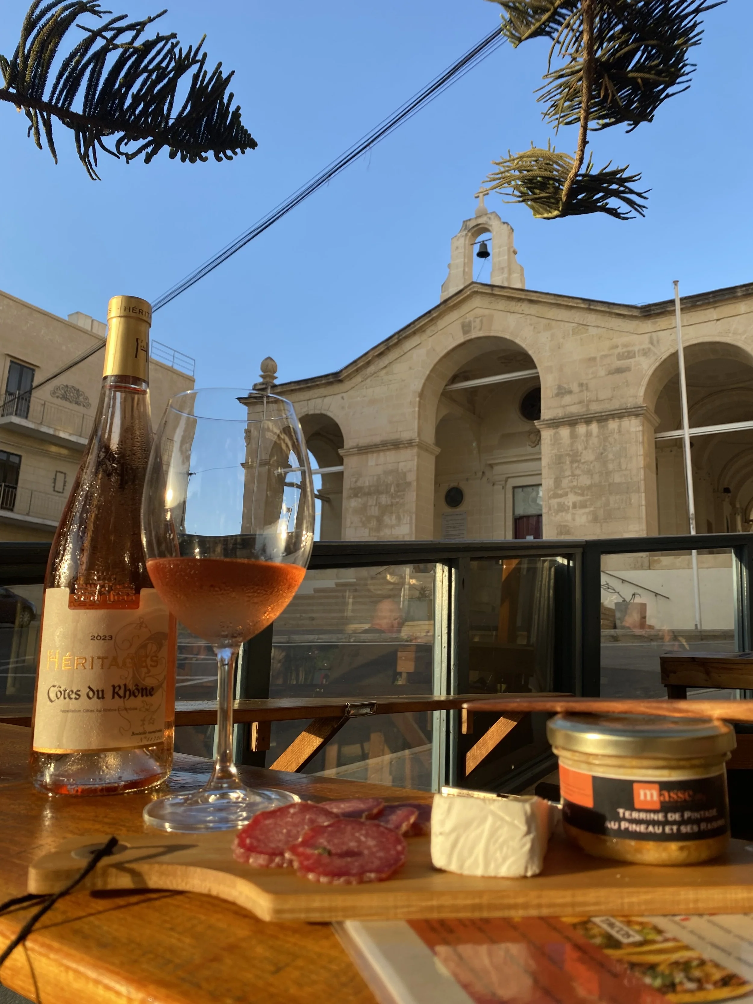 A table setup with a bottle of rosé wine, a glass of rosé wine, slices of cured meat, a piece of cheese, and a jar of pâté, outdoors with a historic stone church in the background under a blue sky.