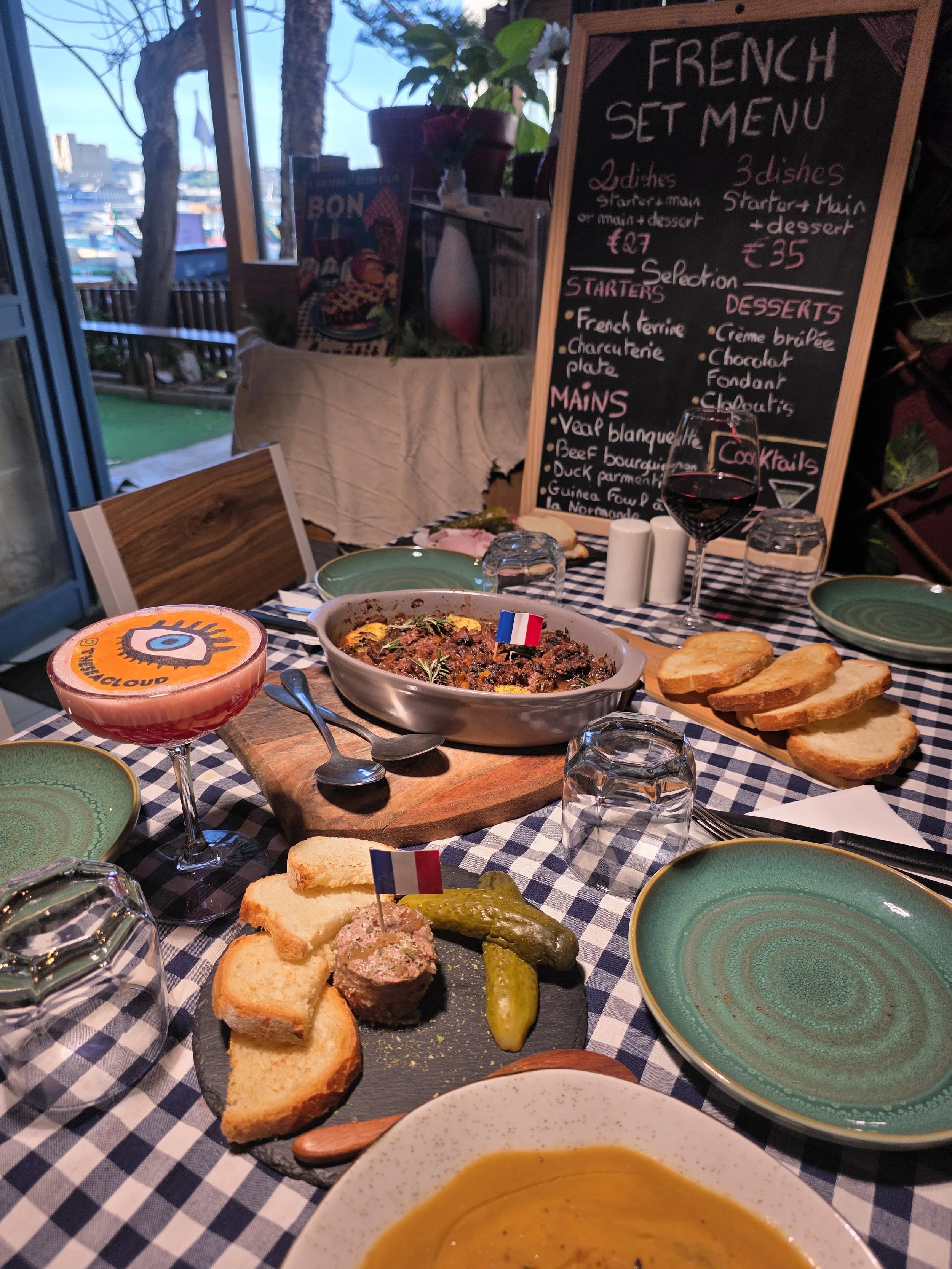 A table set for a French meal with multiple dishes, including bread, a meat dish with a small French flag, pickles, and a colorful drink. In the background, a chalkboard lists a French set menu with options for starters, mains, and desserts. The sett
