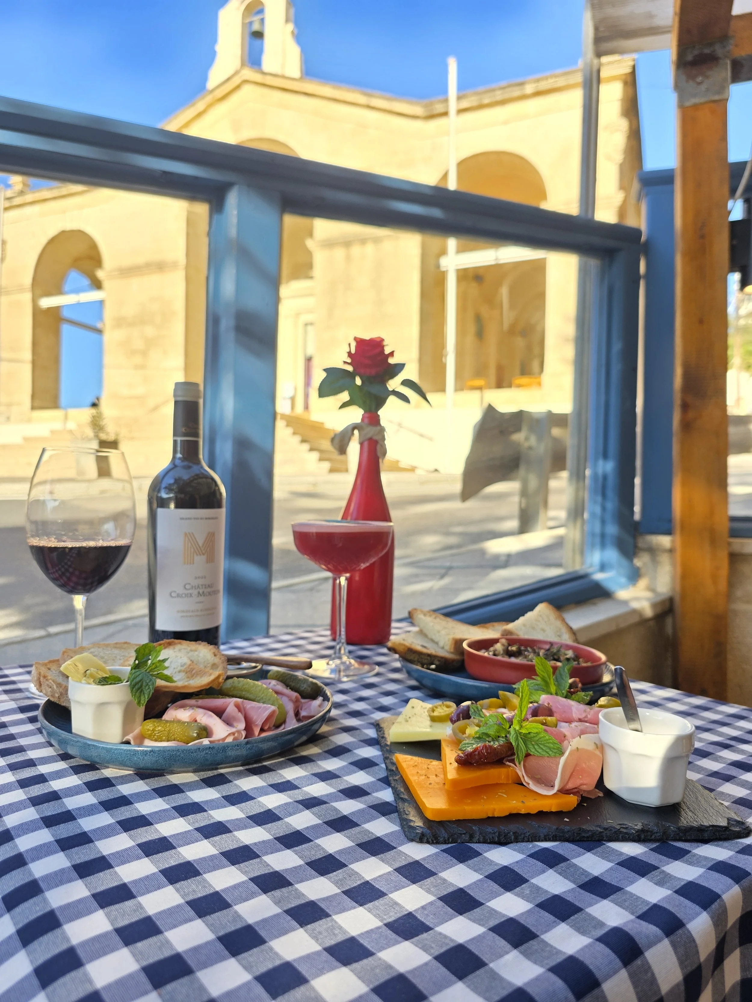 A table set with cheese, meats, bread, a glass of red wine, a bottle of wine, and a cocktail, with a church building in the background.