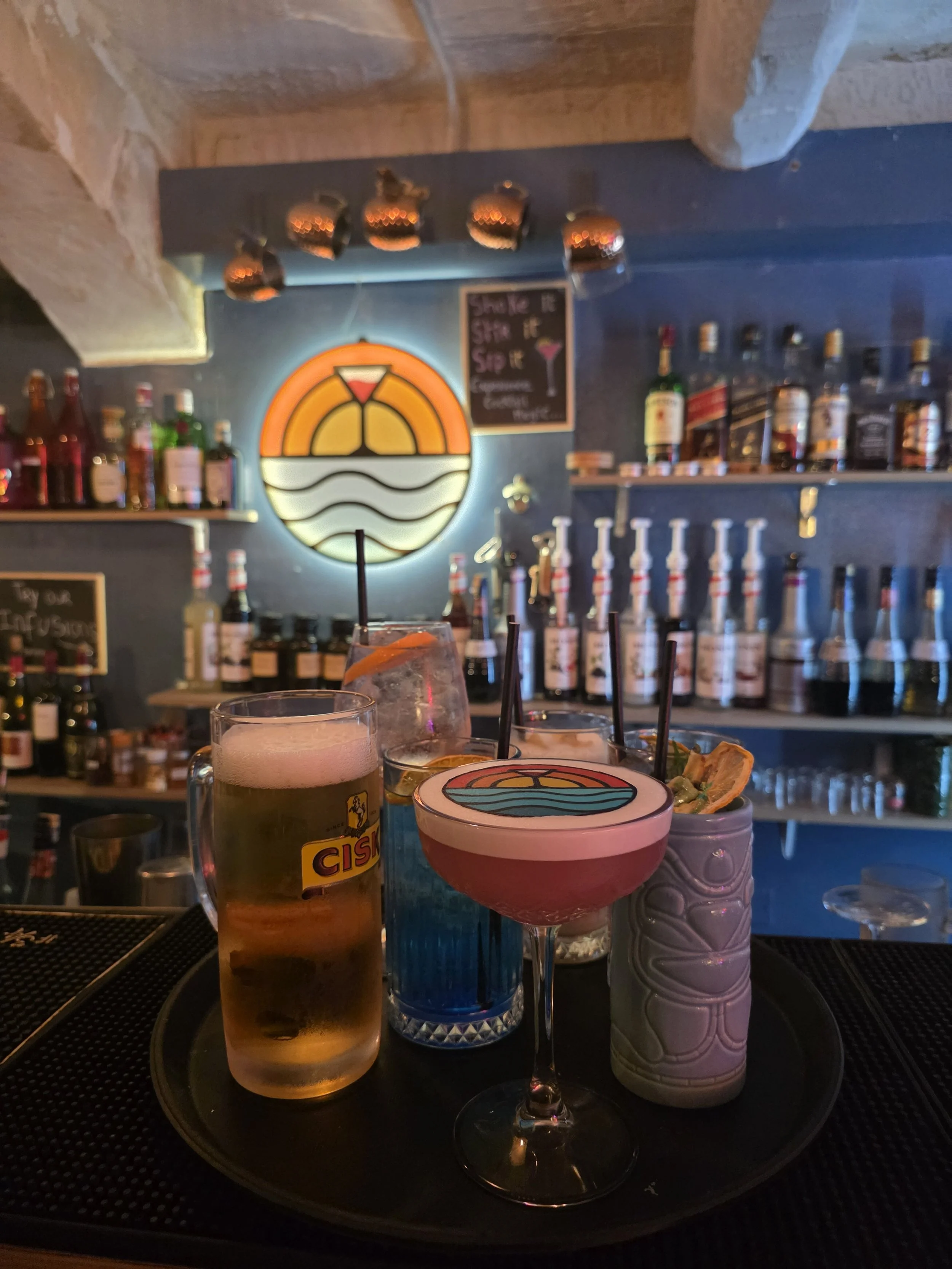 Assorted colorful cocktails on a tray at a bar, with bottles of alcohol on shelves and a neon sunset sign in the background.