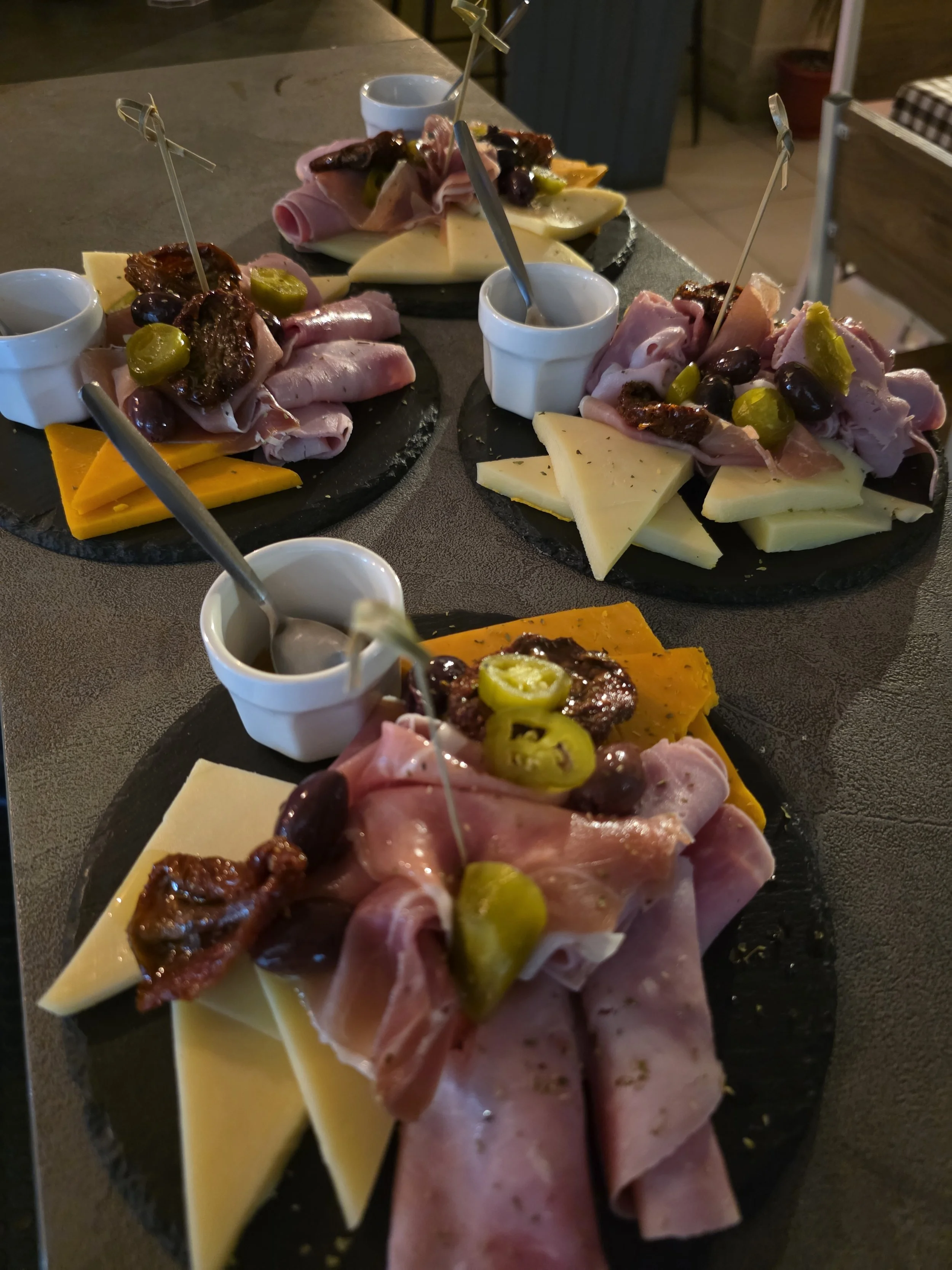 Three charcuterie boards with assorted cheeses, cured meats, olives, sun-dried tomatoes, and small bowls of dipping sauces on a dark table.