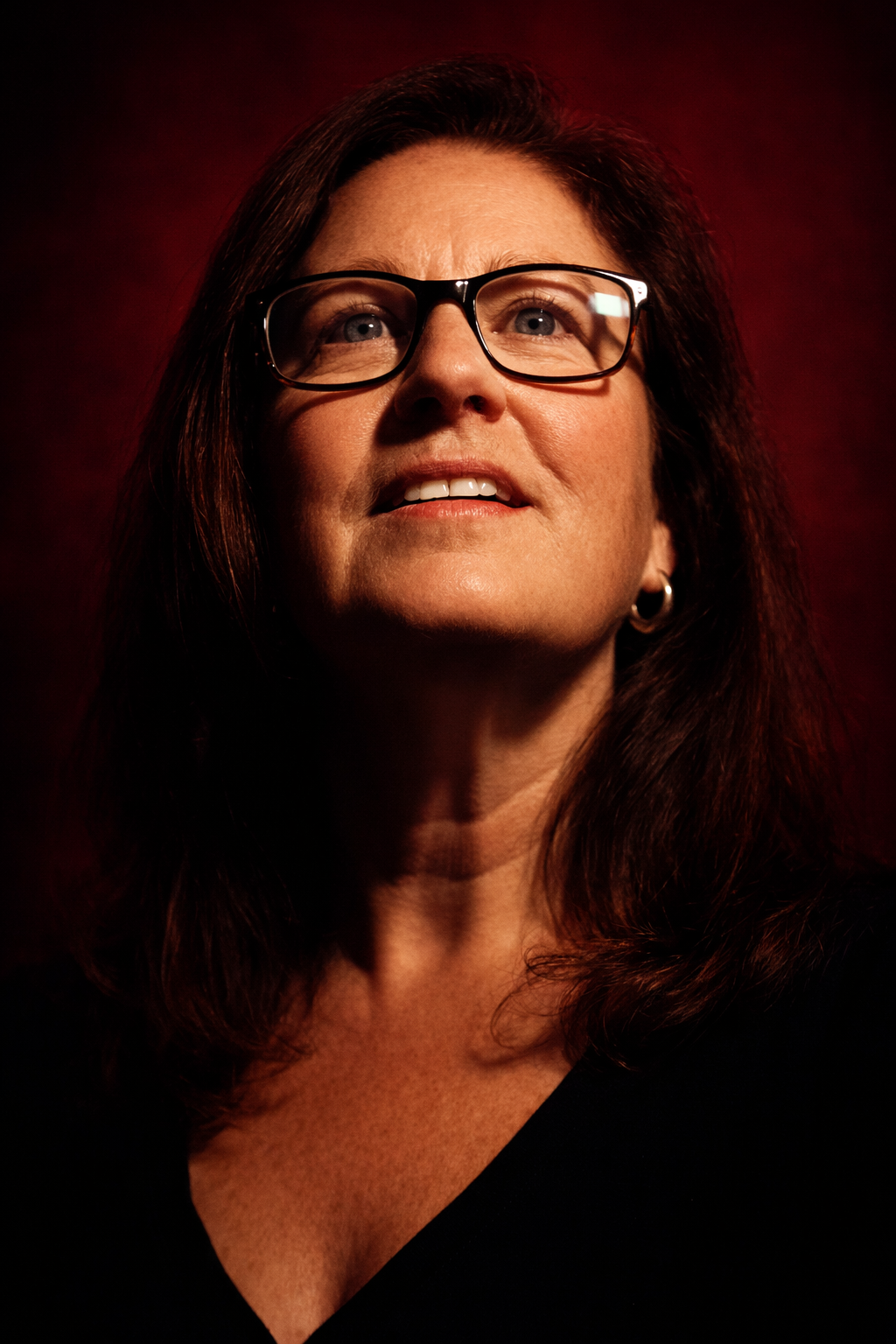Close-up portrait of Renee, wearing glasses, looking slightly upward with a faint smile, against a dark red background.