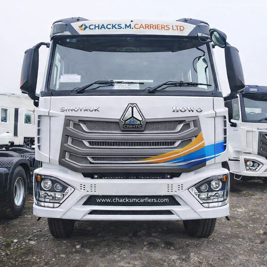 White truck with the words 'Chacks M. Carriers LTD' on top, logo in the center, and website 'www.chacksmcarriers.com' below, parked among other white trucks on gravel lot.