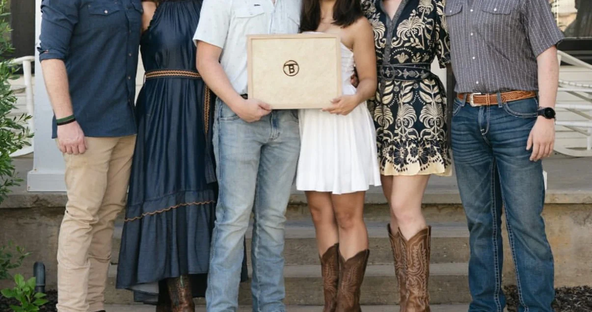 Group of six people standing on steps, holding a wooden box, with plants and a building in the background.