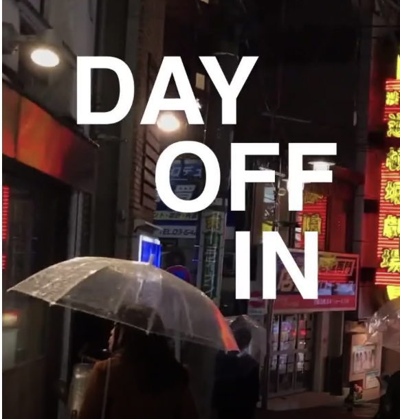 A nighttime street scene with people holding umbrellas in a city with illuminated signs and storefronts. Overlaid text says, 'DAY OFF IN'.