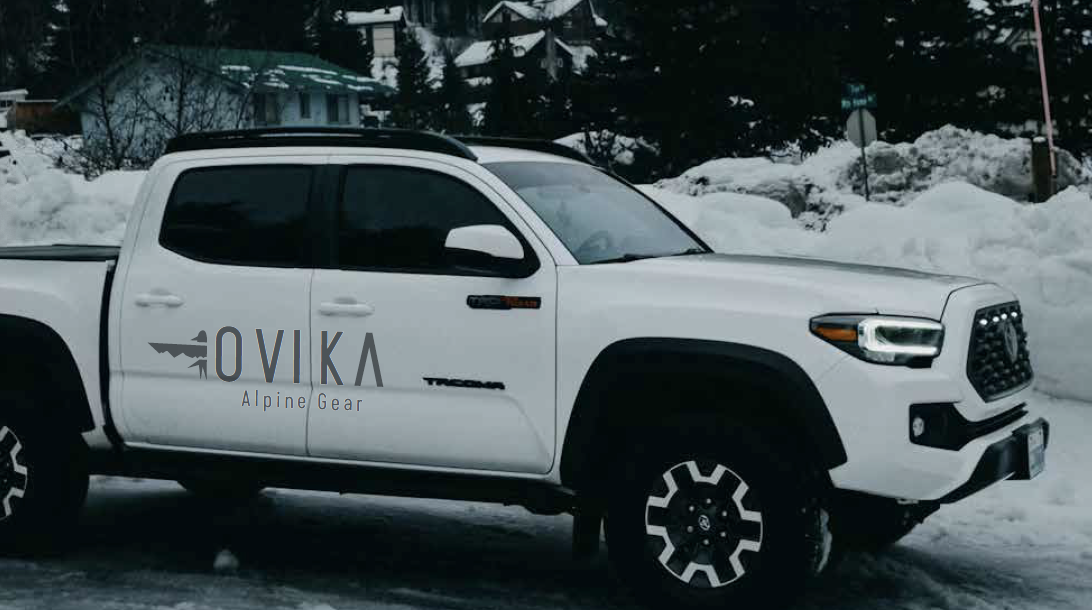 White pickup truck parked on snow-covered ground, with a logo and text on the door reading "OVIKA Alpine Gear," and houses with snow in the background.