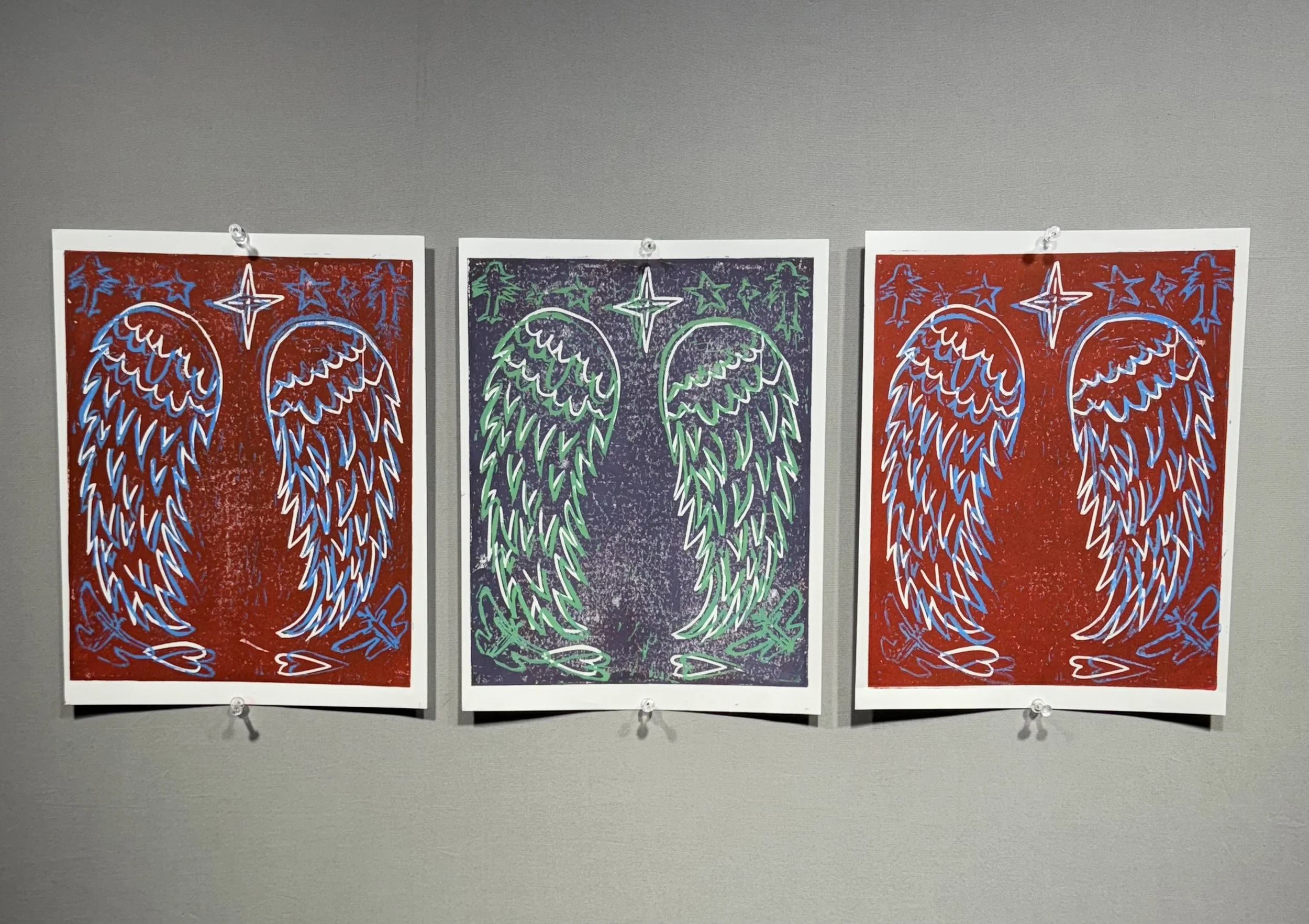 Three posters with angel wings and star designs, arranged in a row on a gray wall.