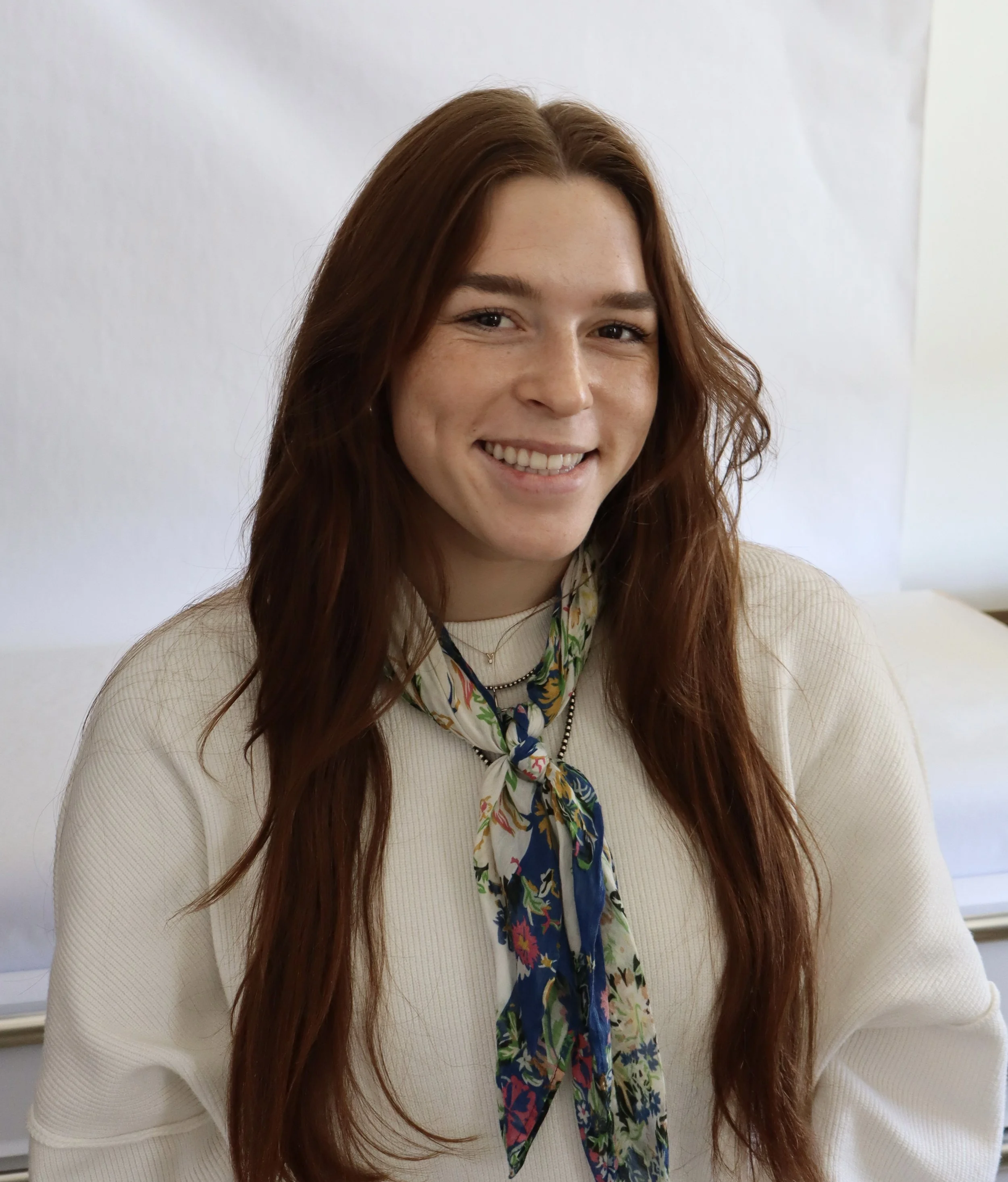 A young woman with long, wavy red hair smiling, wearing a cream-colored sweater and a colorful floral scarf, sitting in a room with white walls.