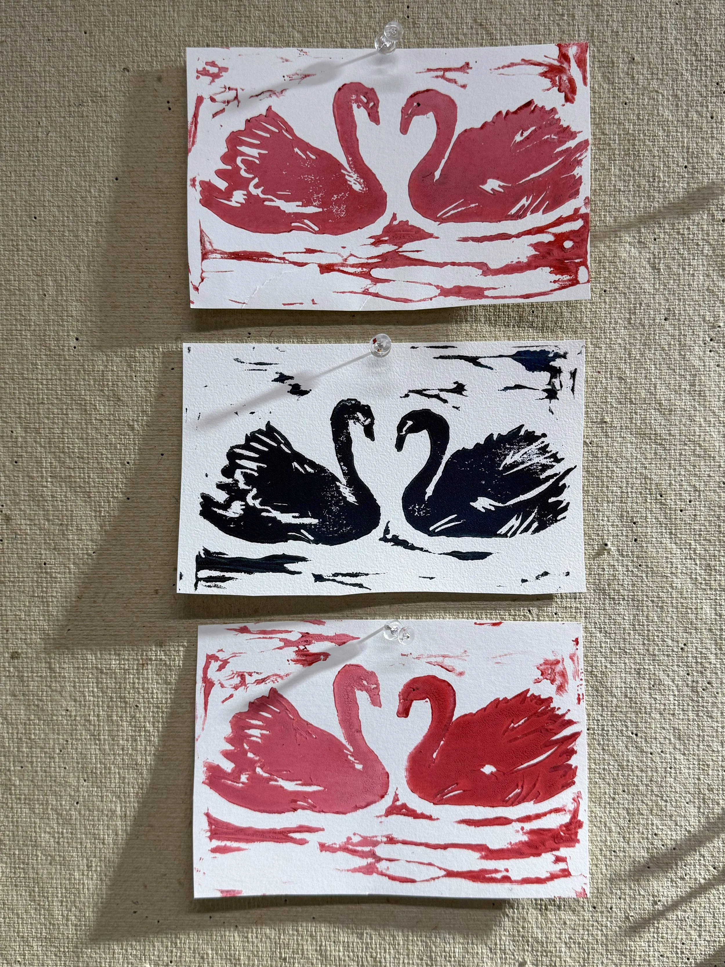 Three pieces of art depicting two swans facing each other on water; the top and bottom are red ink prints, and the middle is a black ink print.