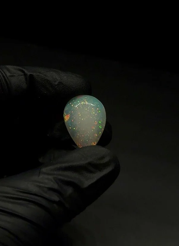 A hand in a black glove holding a pear-shaped opal with colorful iridescence against a dark background.