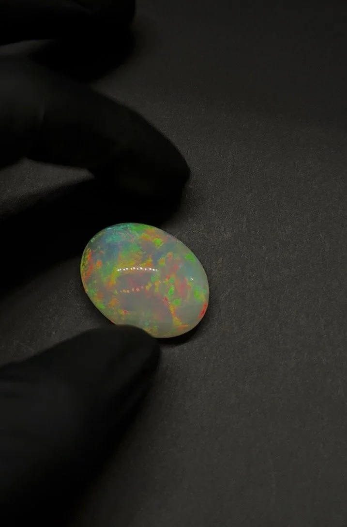 A gloved hand holding an oval-shaped opal gemstone with colorful play-of-color against a black background.