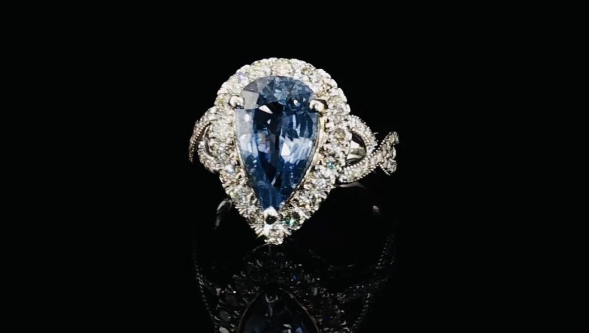 A silver ring with a large pear-shaped blue gemstone surrounded by small diamonds, set against a black background.