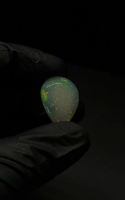 A gloved hand holding a glowing opal gemstone with green, yellow, and orange patterns against a black background.