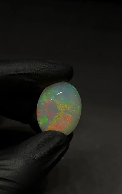 A person wearing black gloves holding a glowing opal gemstone with colorful internal play-of-color against a dark background.