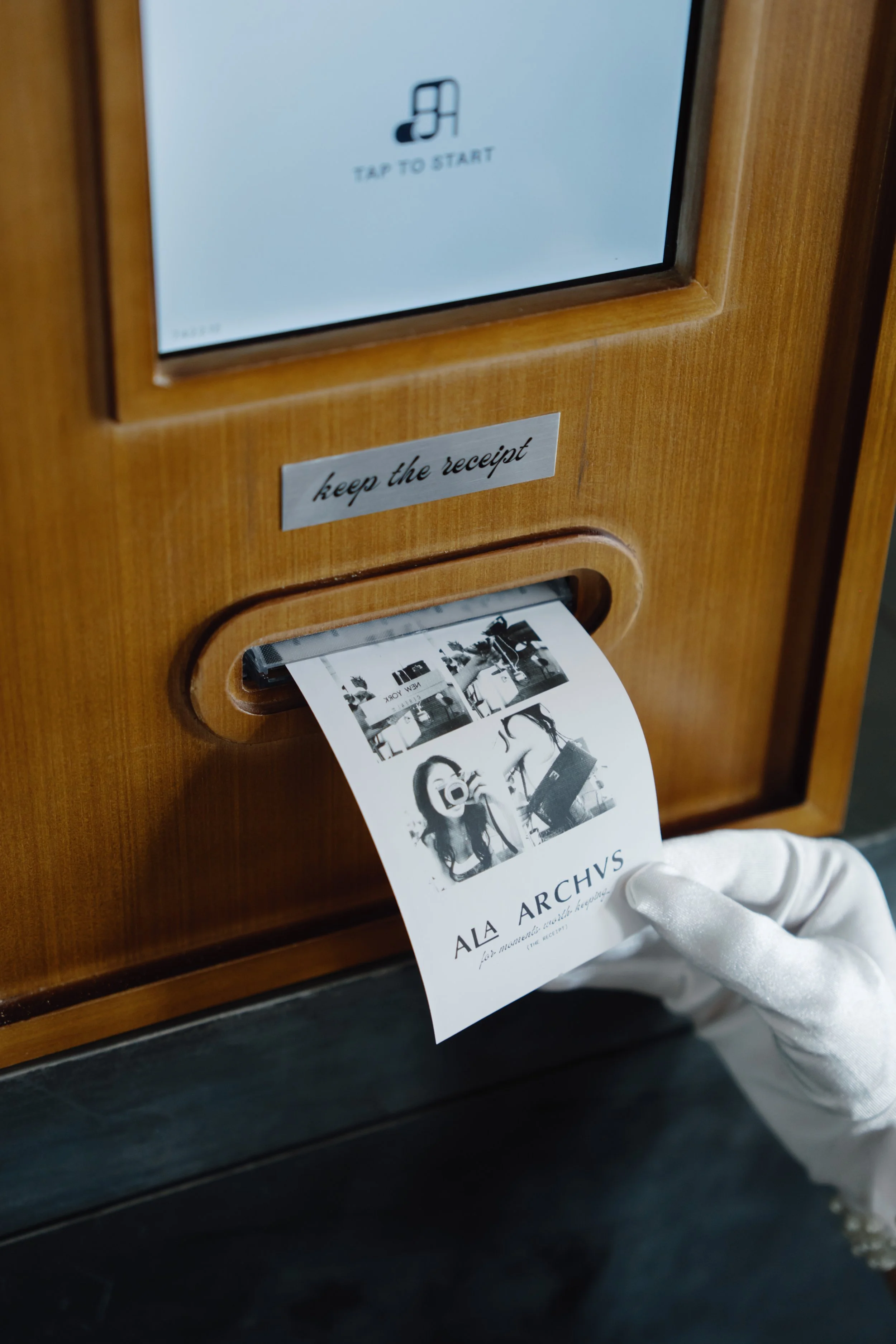 A person wearing a white glove is inserting a black and white photo booth printout into a wooden kiosk with a screen that says 'Tap to Start' and a metal plaque that says 'keep the receipt'.