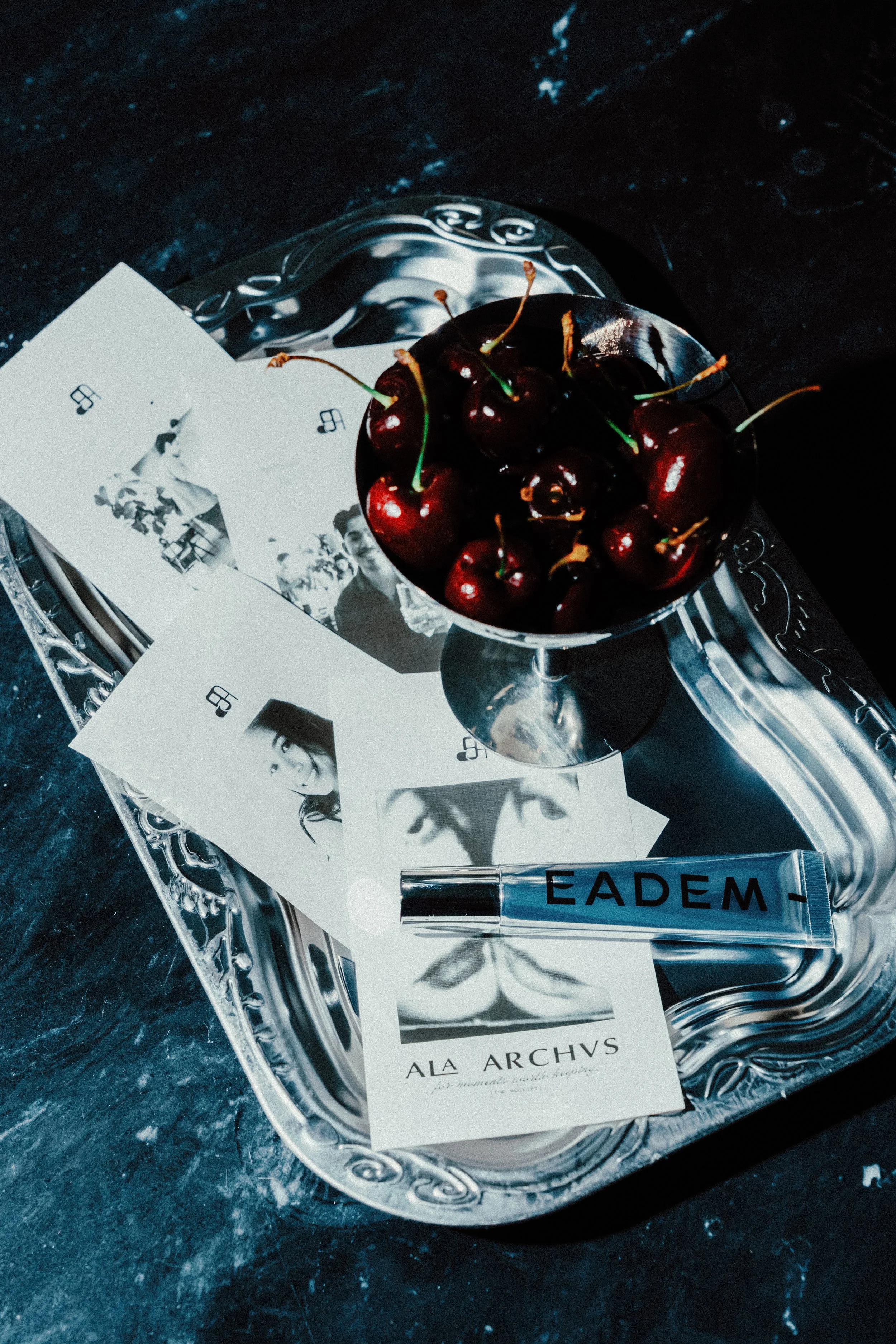 A silver tray holding a small bowl of cherries, black and white photographs of people, a lipstick tube labeled 'EADEM', and some printed text on a dark surface.