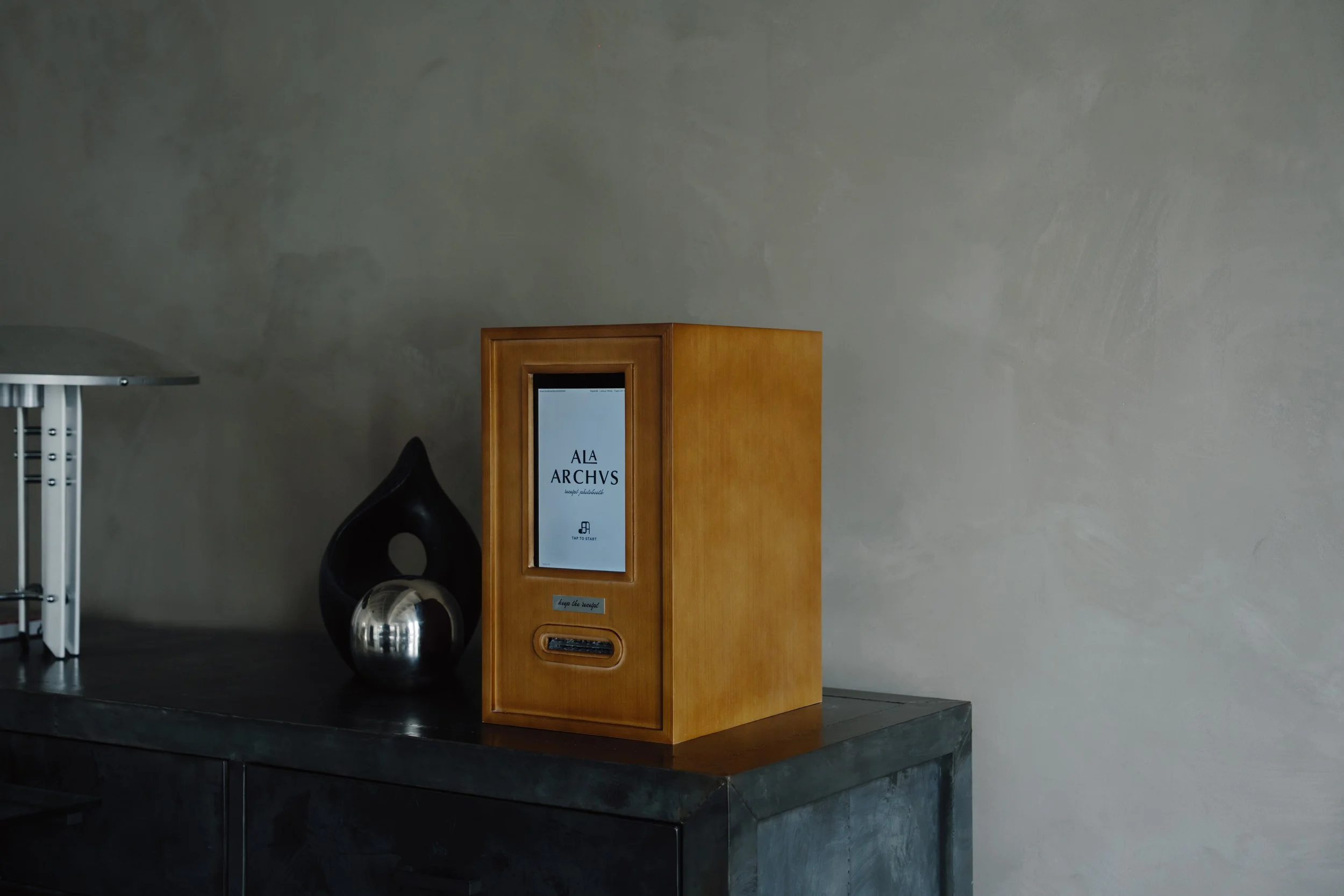 A wooden display stand with a touchscreen showing an app named 'Ala Archivs' on a dark gray surface, with a black decorative sculpture and a small metallic object beside it, against a plain gray wall.