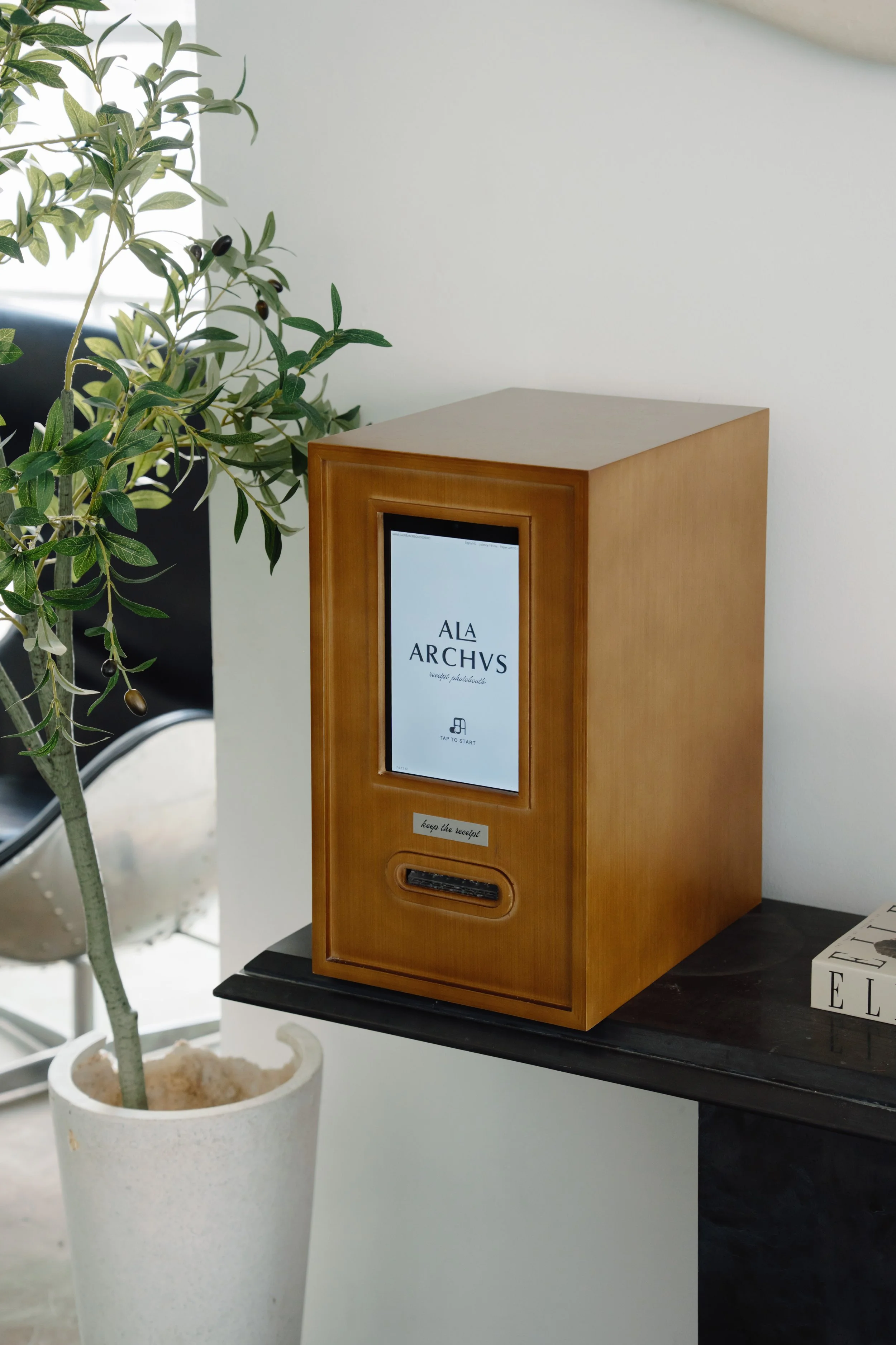 A wooden receipt kiosk with a digital screen displaying the words "ALARCHVS," placed on a black table near a potted plant and a book.