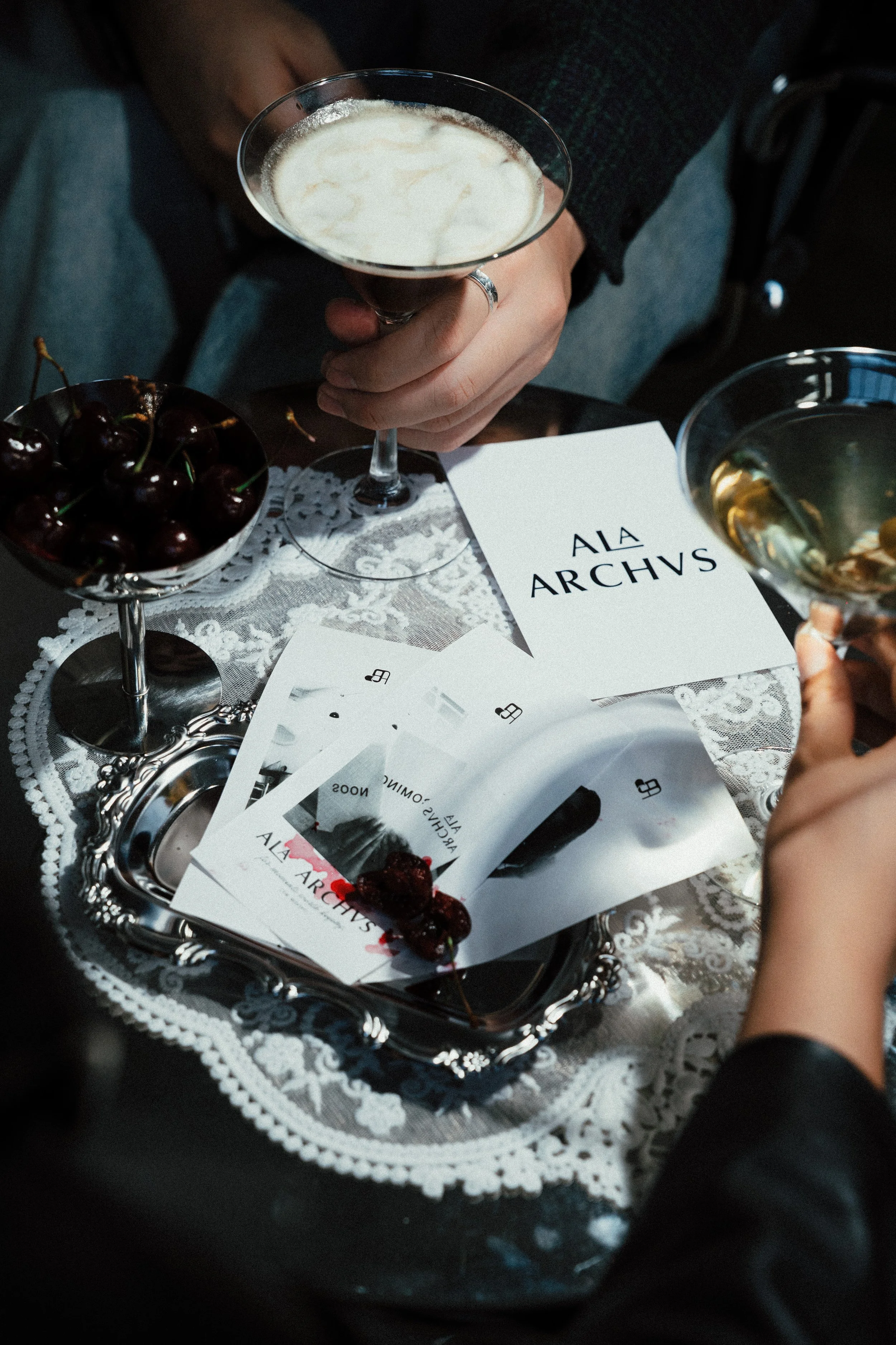 Glamorous table setting with two drinks, a bowl of cherries, and printed papers on a lace tablecloth, with a person holding a cocktail glass.