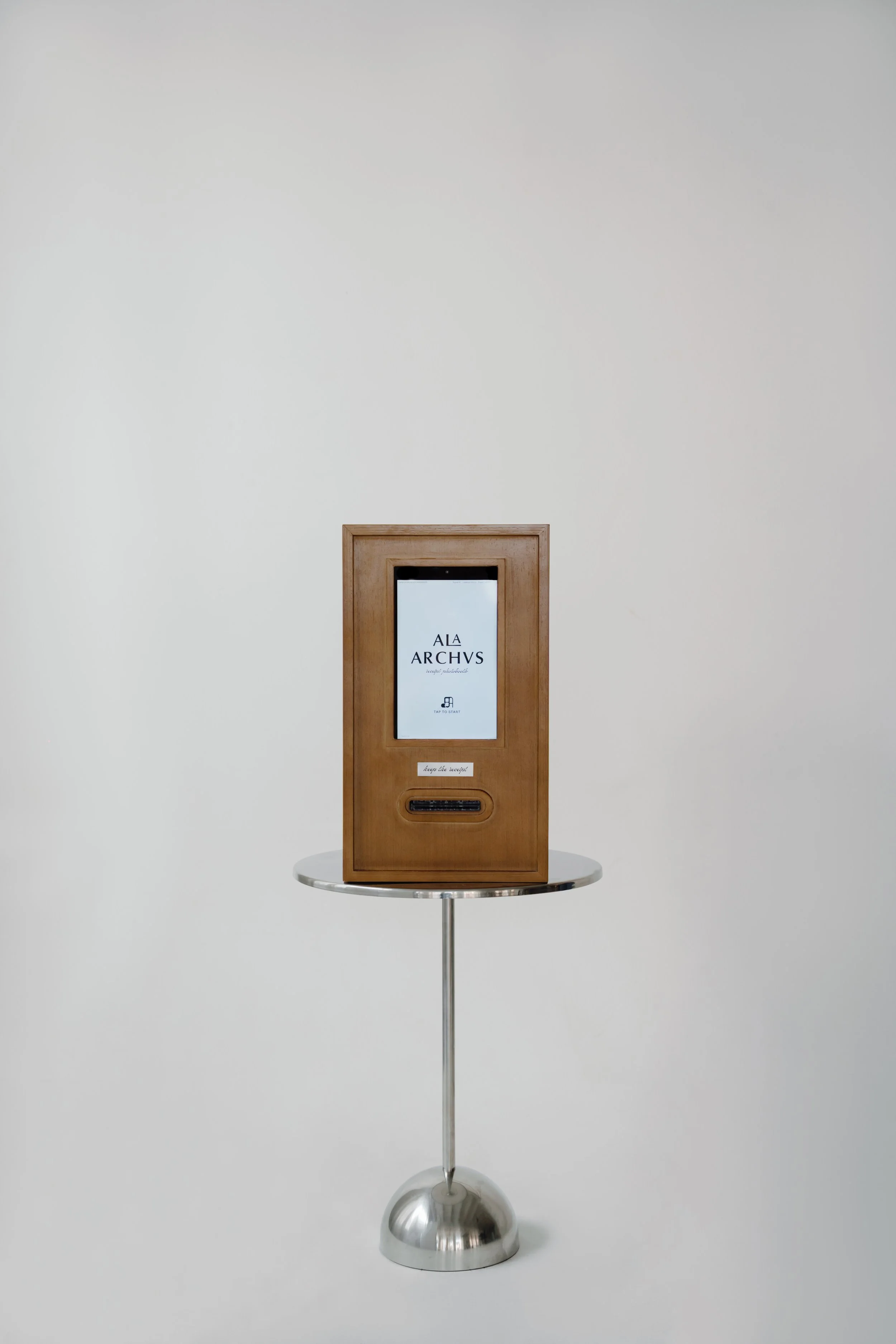 A wooden digital kiosk with a screen displaying 'A LA ARCHIVS' and a small button underneath, placed on a round metal stand against a plain white wall.