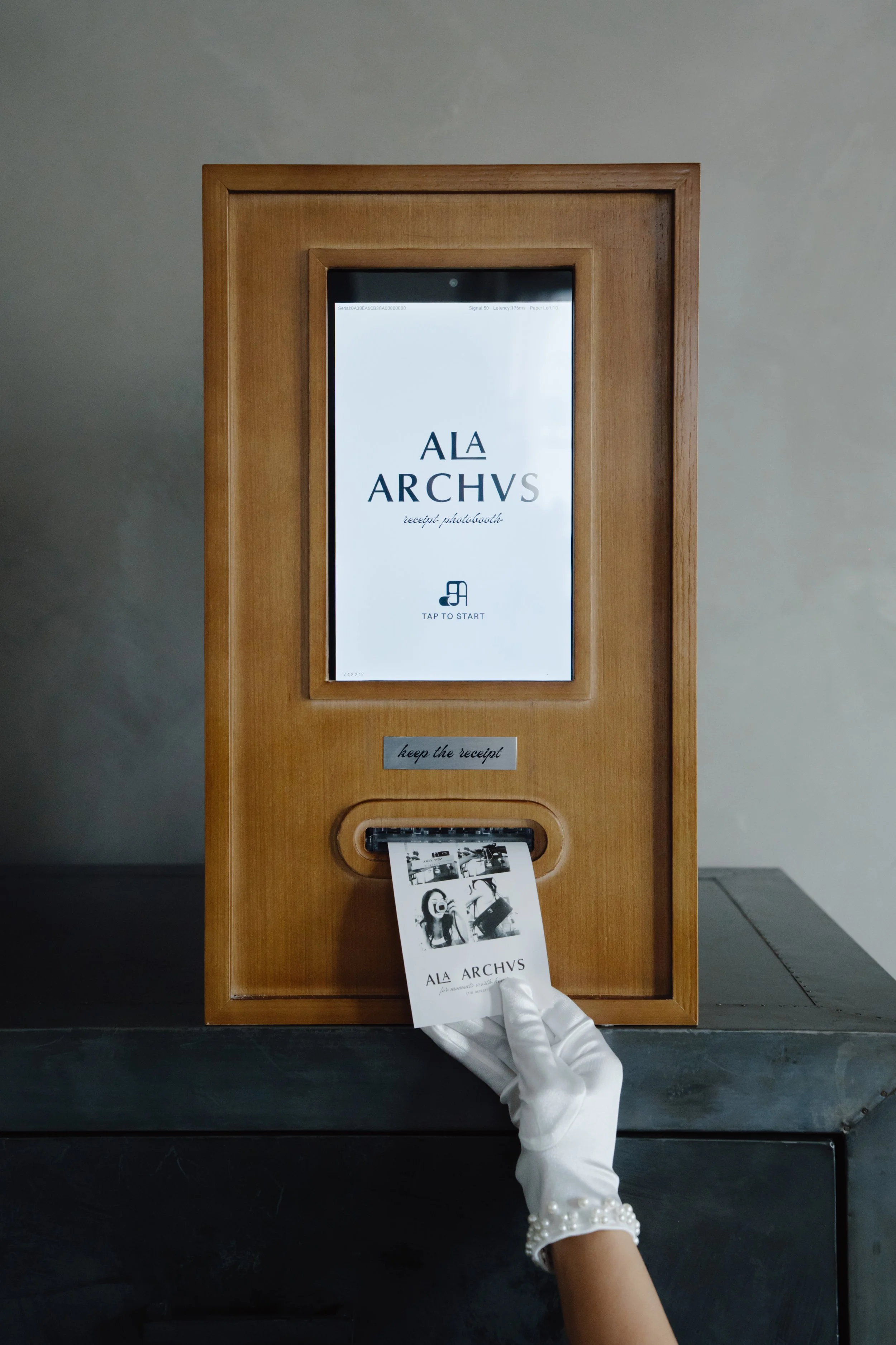 A person wearing a white glove is inserting a photo card into a photo kiosk with a wooden frame, displaying the words 'A LA ARCHVS' and instructions to receive photos and keep the receipt.