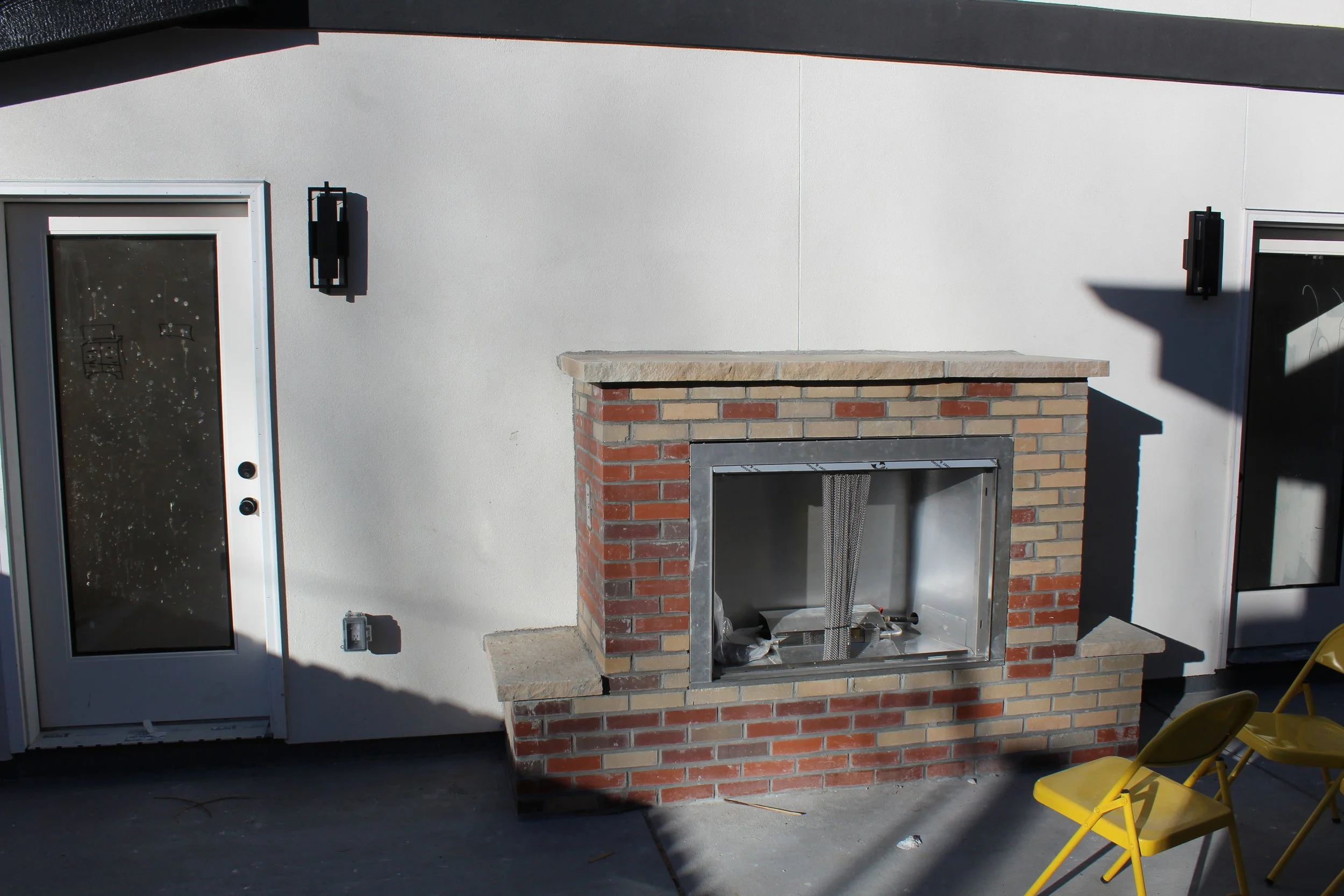 Exterior of a white house with a new brick fireplace and two outdoor yellow chairs. There are two black outdoor lights, one on each side of a glass door, with a smaller metallic switch on the wall below the left light. A professional and super detail