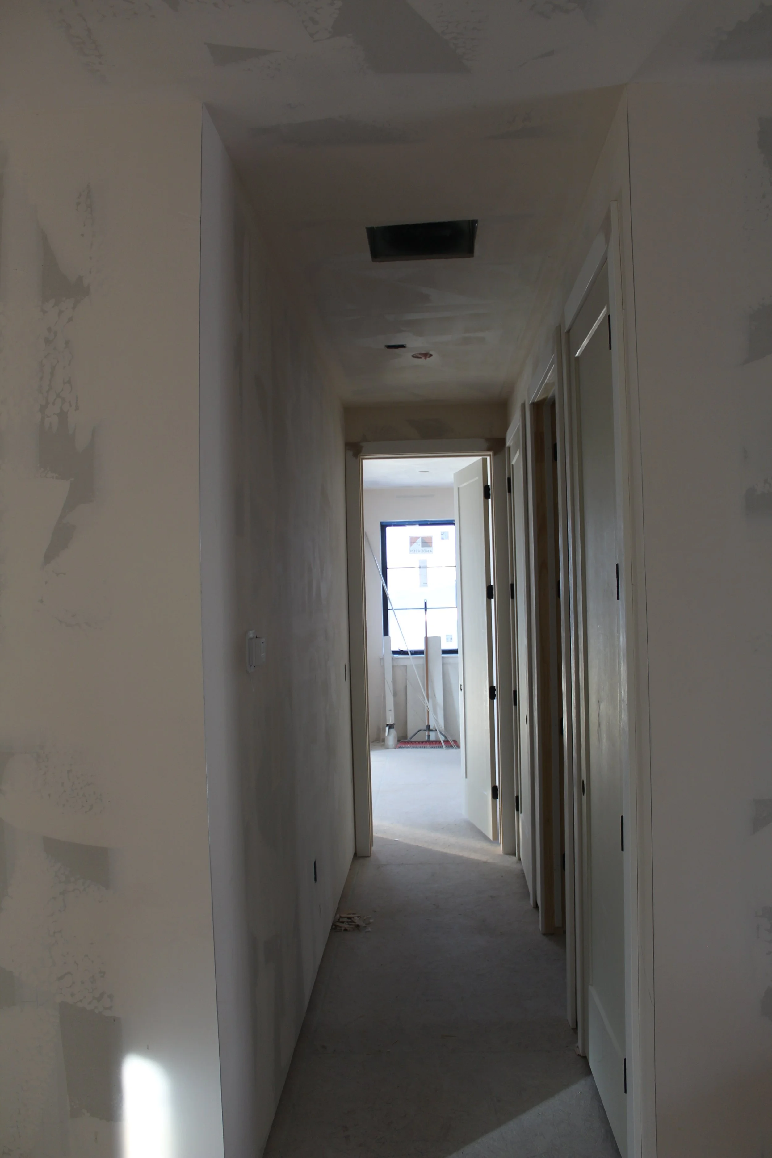A hallway under construction with walls partially painted, an open door at the end leading to a room with a window, and construction equipment near the window.