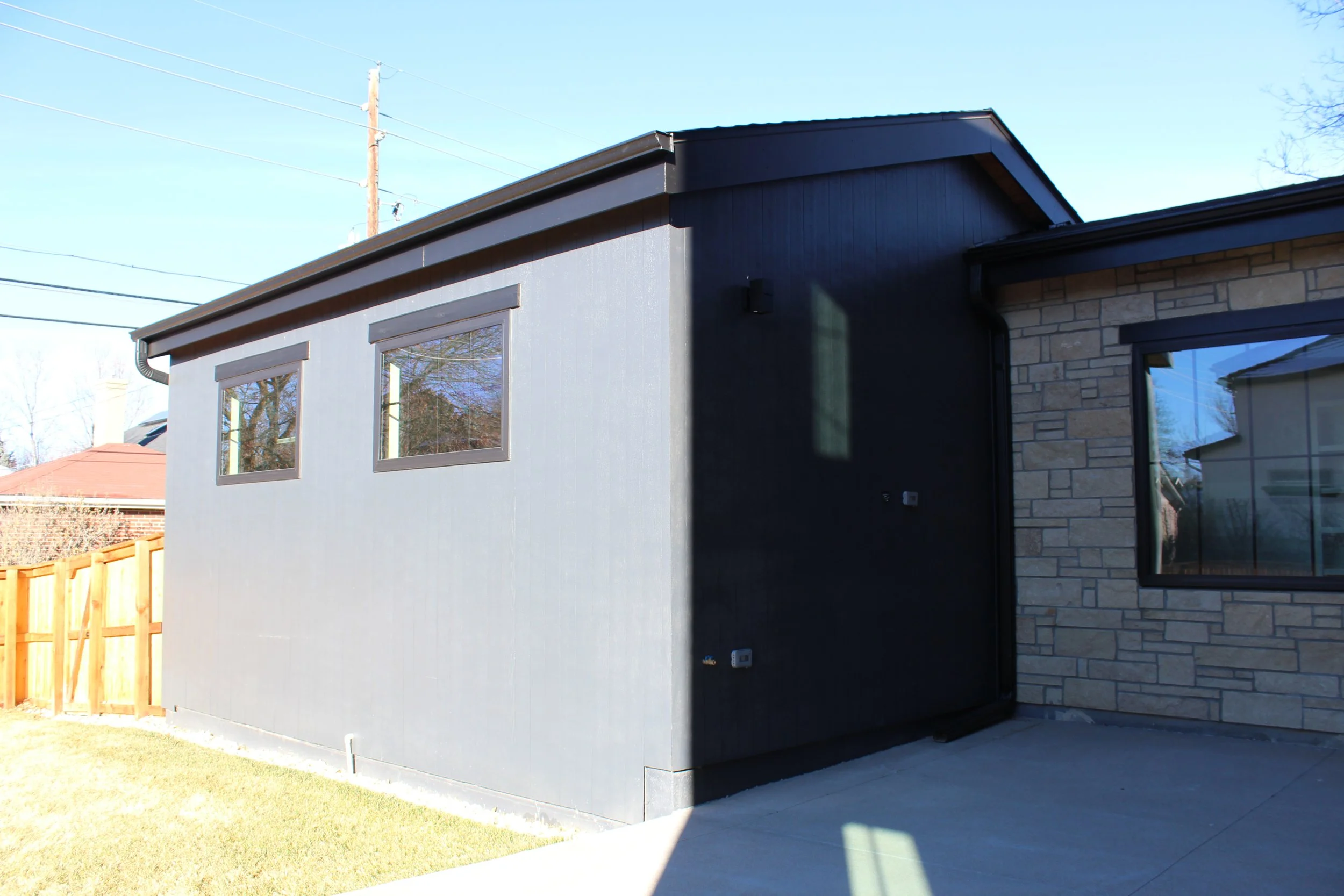 A modern house exterior with two-toned walls, one part painted dark gray and the other part covered with beige brick. There are two windows on the gray part reflecting trees and sky, a concrete patio, a wooden fence, and power lines overhead.