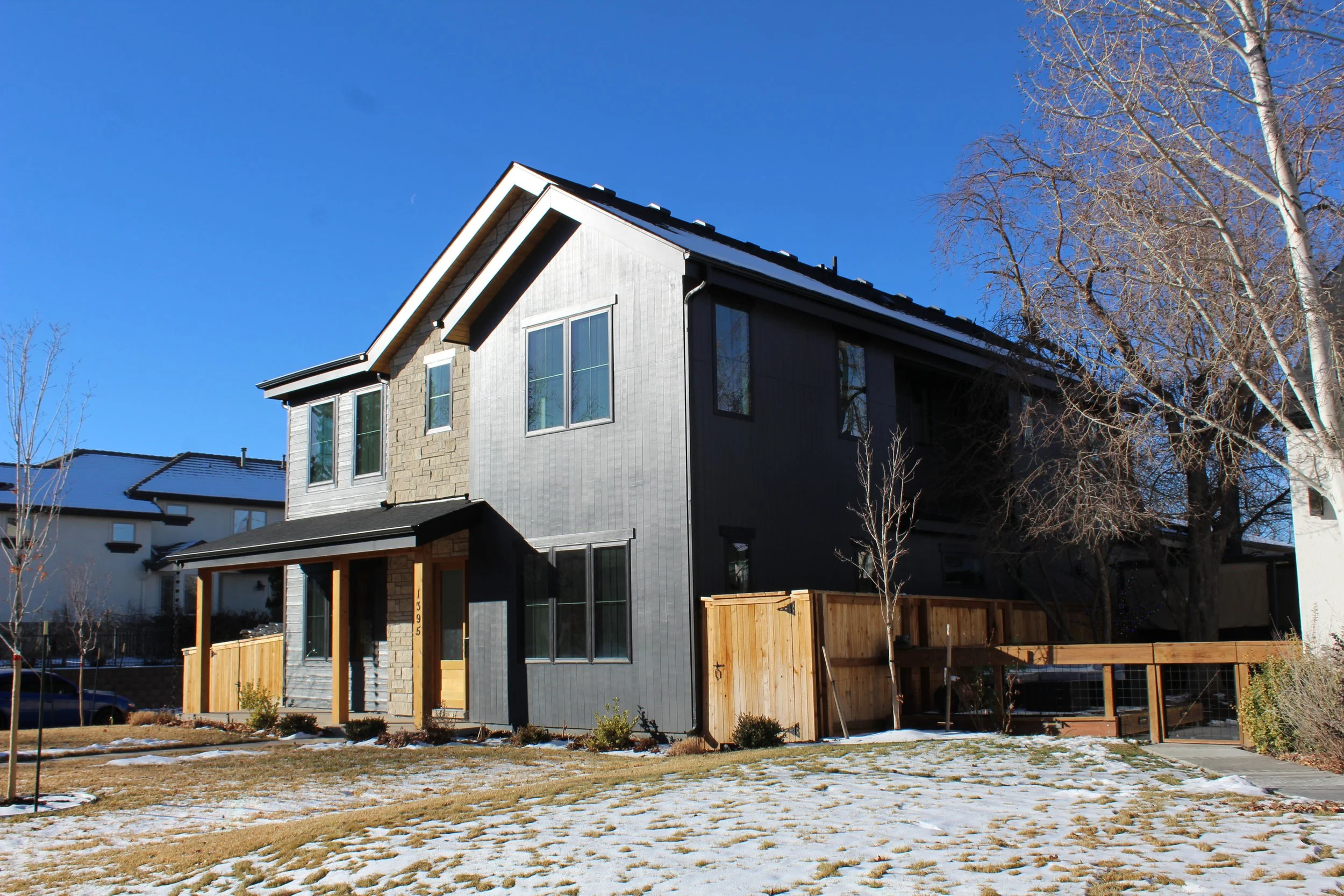 Modern two-story house with a mix of dark siding and stone exterior, front porch, and snow-dusted lawn, under a clear blue sky.