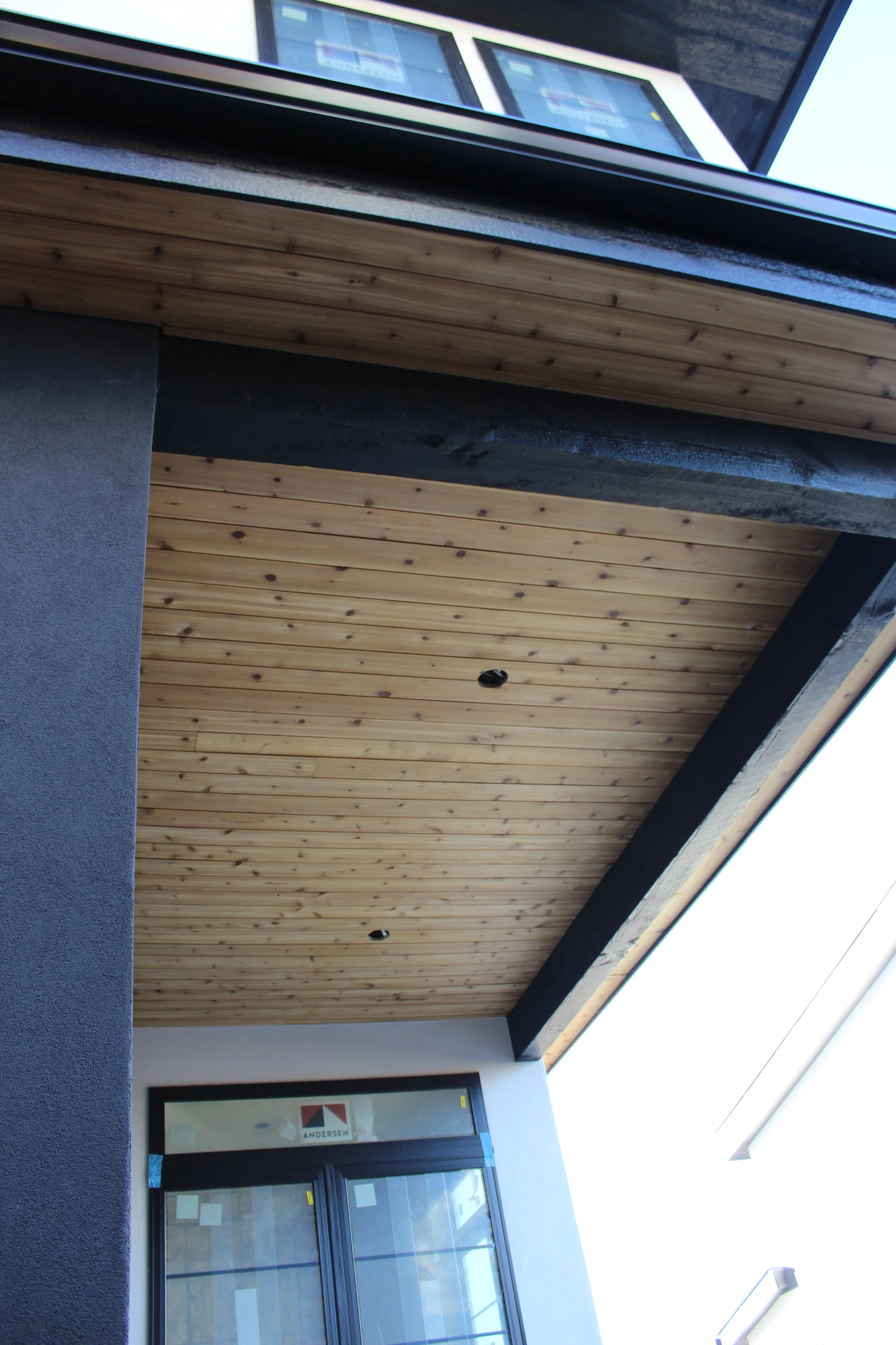 Close-up of the ceiling of a building's entrance with wooden panels and black beams, and part of a window and wall. A professional and super detailed work with premium materials. This is a custom house constructed by Modern Traditions Inc     