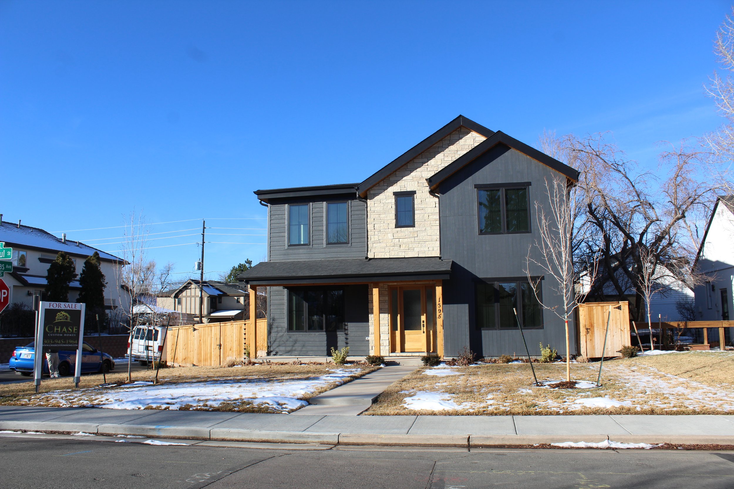 New two-story house with black and beige exterior, large front windows, wooden front door, small front yard with patches of snow, and wooden fence, in a suburban neighborhood.