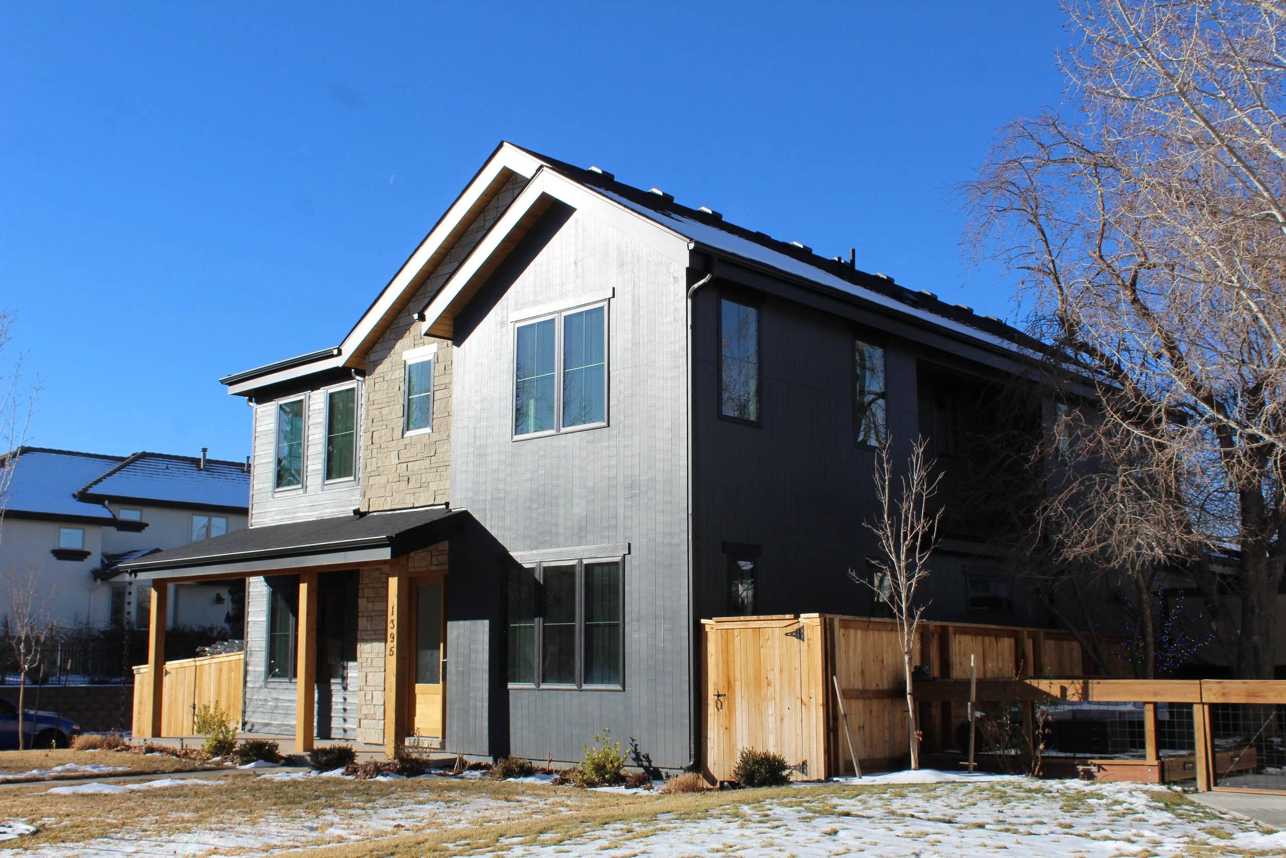 Modern multi-story house with a grey exterior, stone and wood accents, and a fenced yard with patches of snow under a clear blue sky.