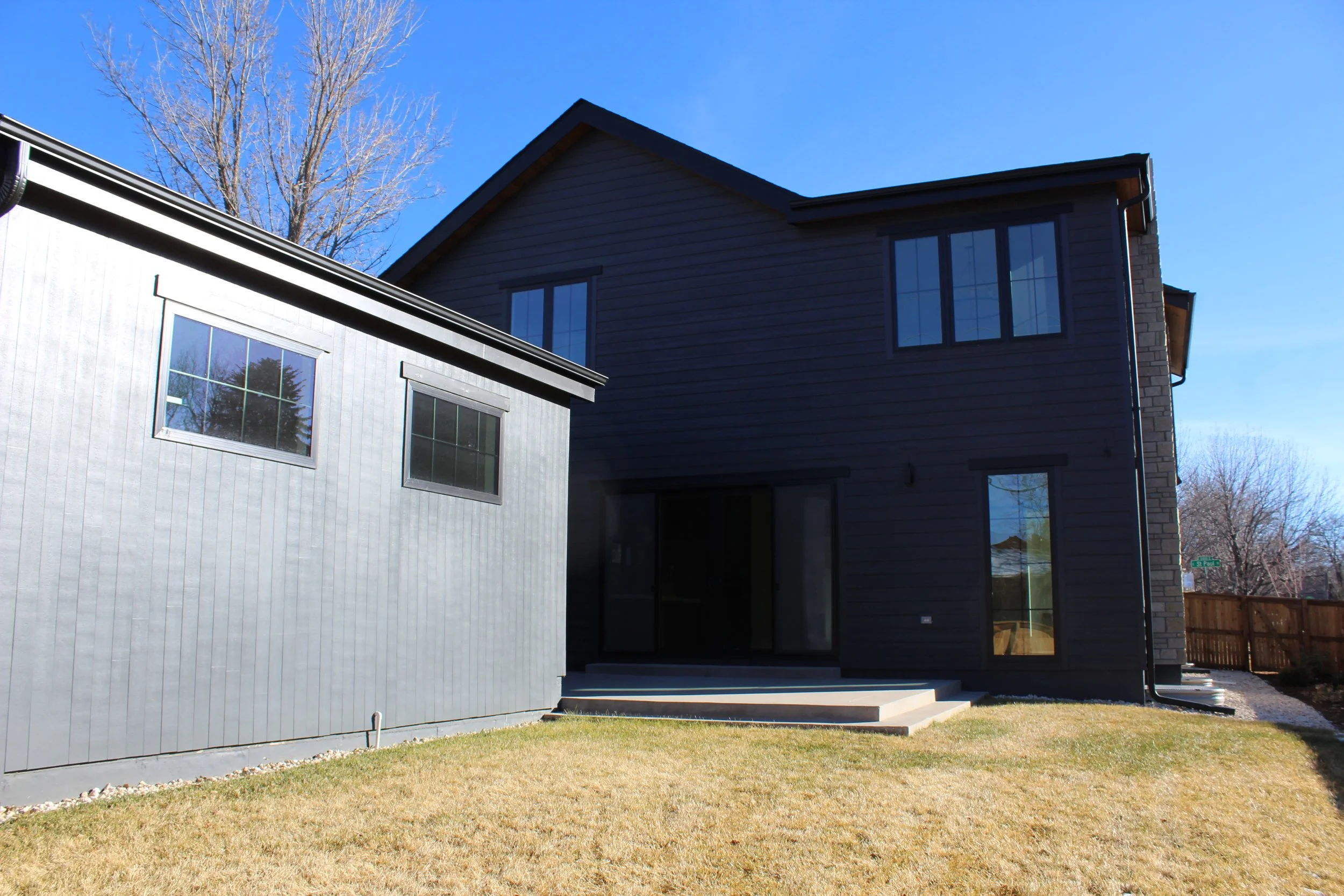 Backyard view of a modern two-story house with black siding, multiple windows, and a small concrete patio with steps on a clear, sunny day.
