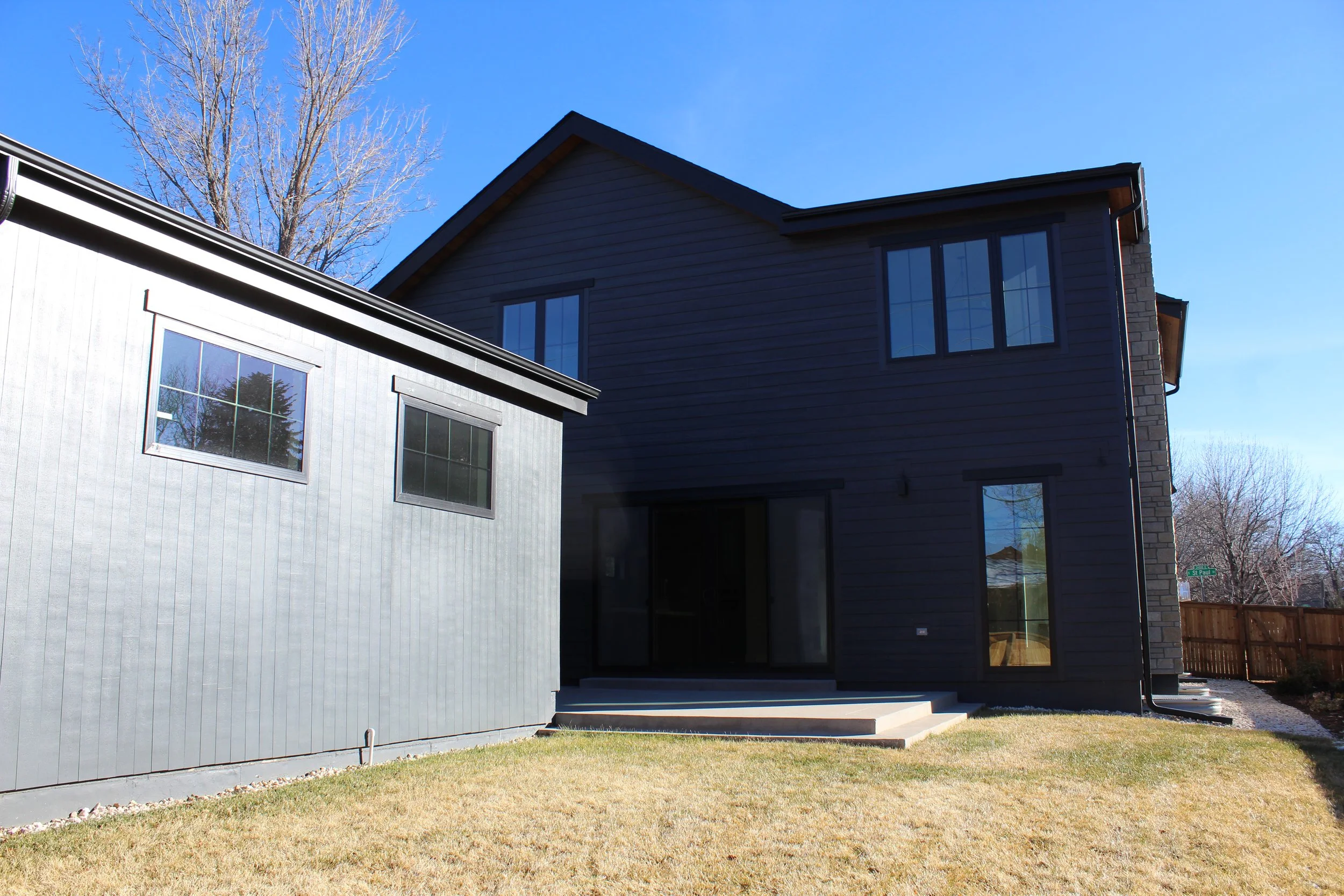 View of a modern two-story house with a dark exterior, large windows, and a small set of steps leading to a sliding glass door. A grassy yard and a wooden fence are visible. Clear blue sky in the background. A professional and super detailed work wit