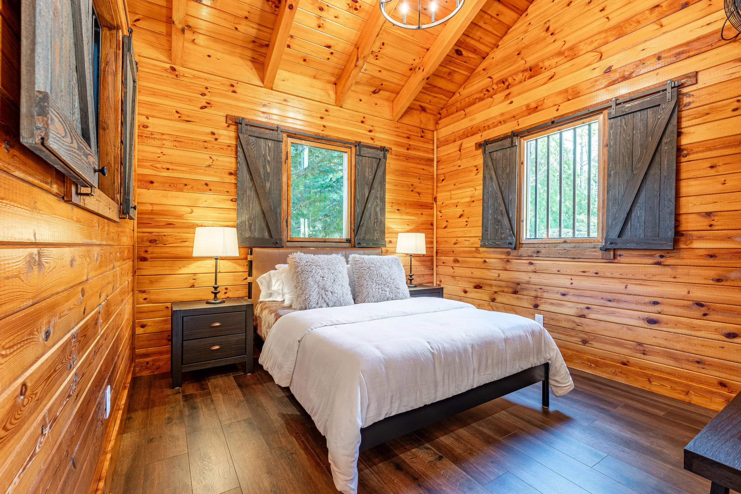 A cozy bedroom with wooden walls, two windows with dark wooden shutters, a bed with white linens and fluffy pillows, two lamps on dark nightstands, and wooden flooring.