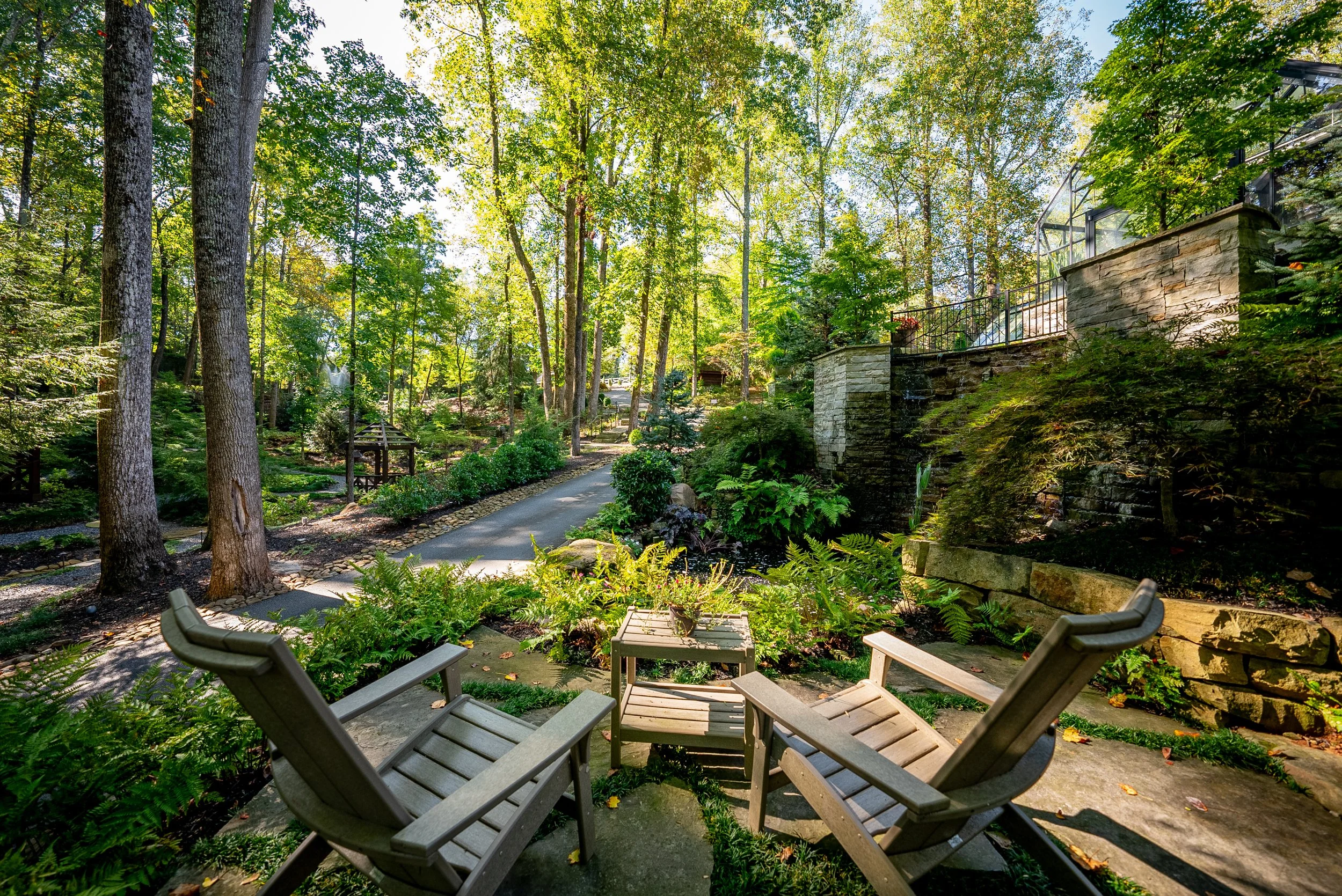 A peaceful backyard garden with two wooden lounge chairs facing each other, a small table, lush green foliage, tall trees, a winding pathway, and a stone wall with a glass porch on top.