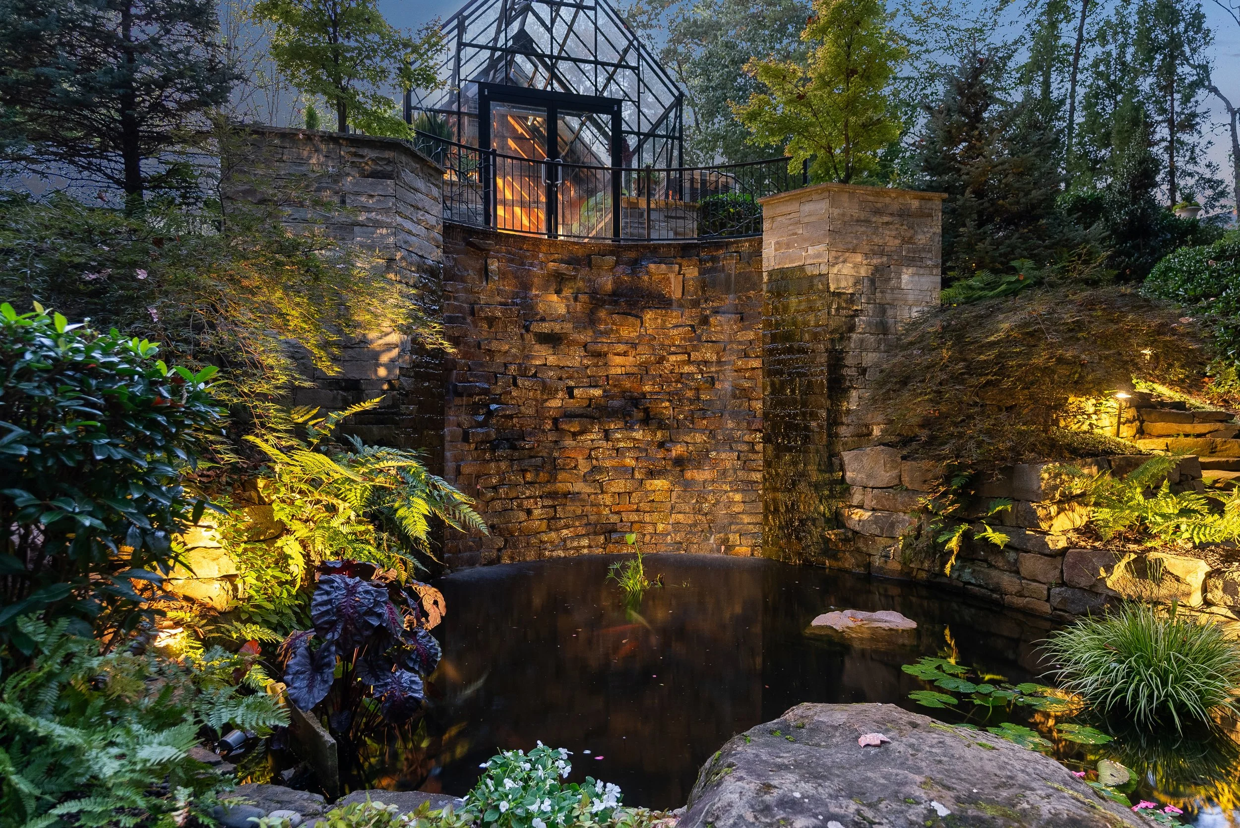 A landscaped garden with a pond, stone waterfall feature, lush greenery, and an illuminated glass greenhouse in the background, during dusk.