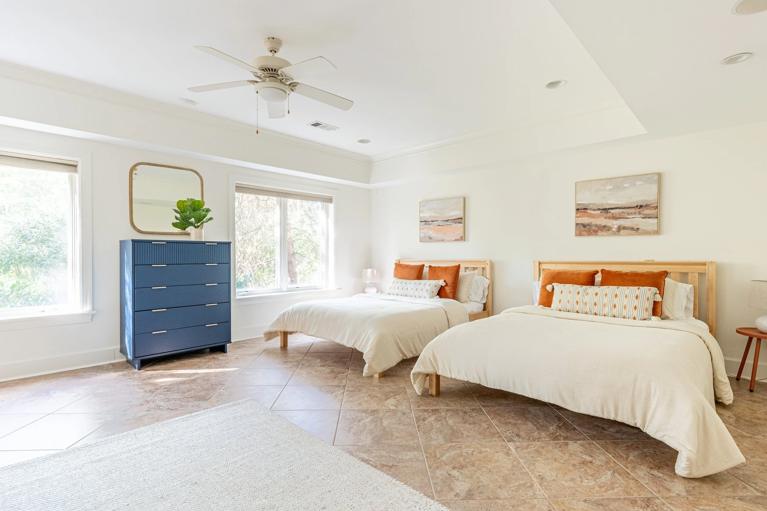 A bright bedroom with two beds, each with beige bedding and orange pillows. Contains a blue dresser, a mirror, a potted plant, and wall art, with sunlight coming through two windows.