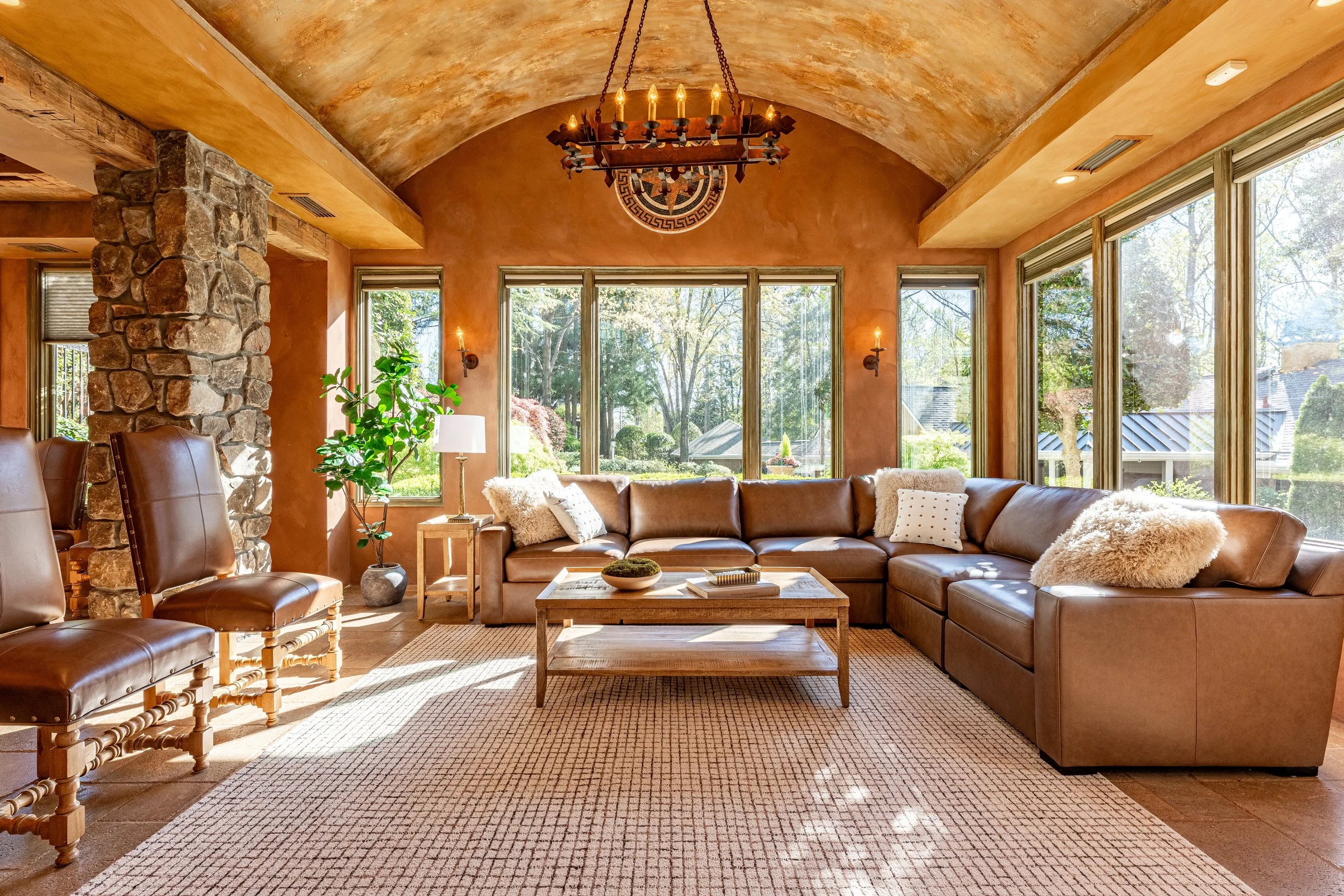 Bright living room with large windows, tan leather sectional sofa, stone pillar, wooden chandelier, potted plant, and outdoor view.