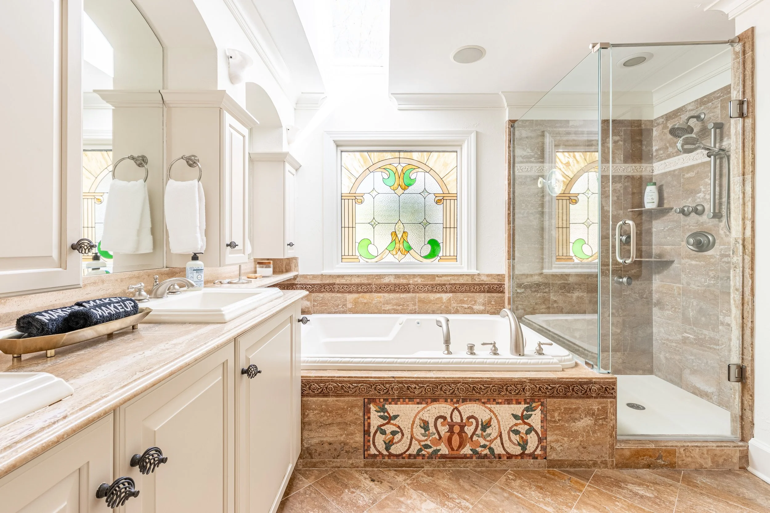 Bathroom with a large bathtub, a glass-enclosed shower, beige and tan marble tiles, white cabinetry, and a stained glass window.