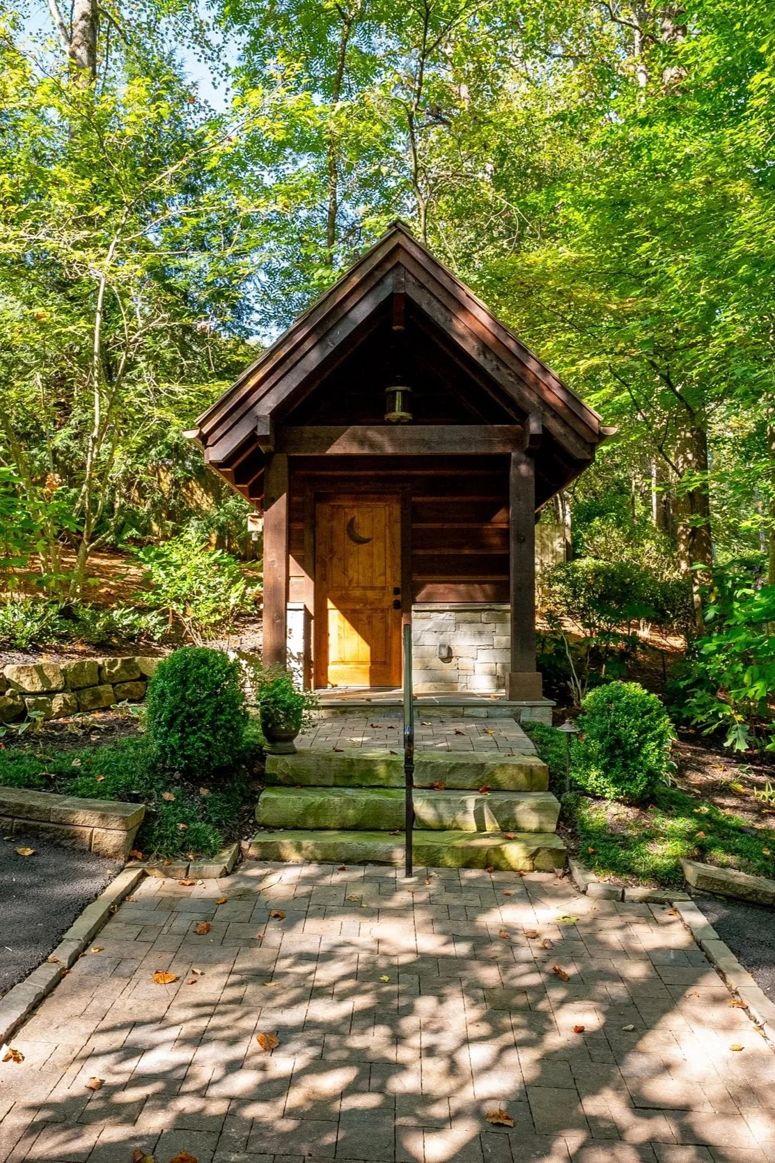 A small wooden cottage with a peaked roof, surrounded by lush green trees, with stone steps leading to a wooden door with a crescent moon carving.