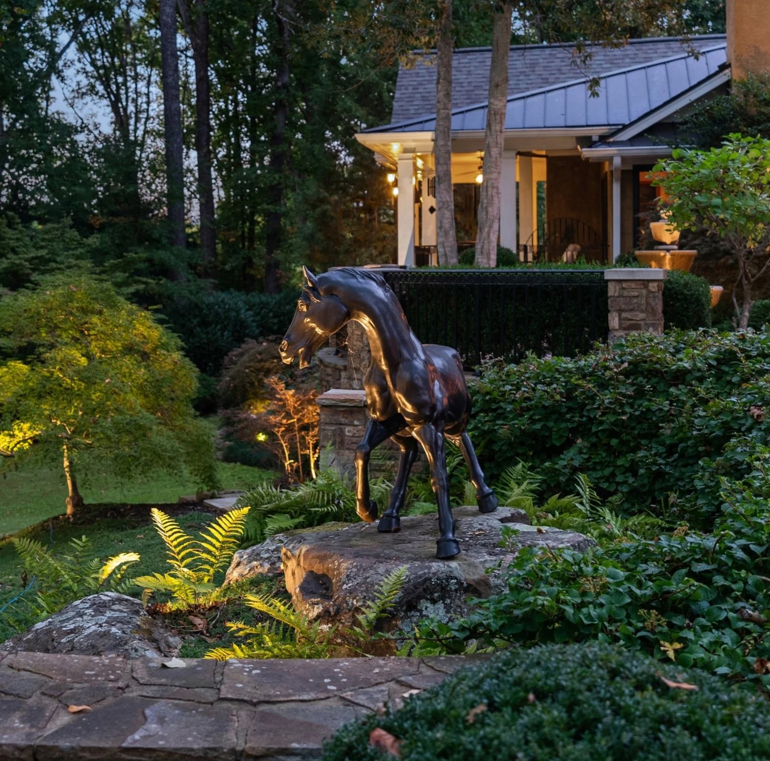 A bronze horse statue in a lush garden at dusk, with a house in the background.