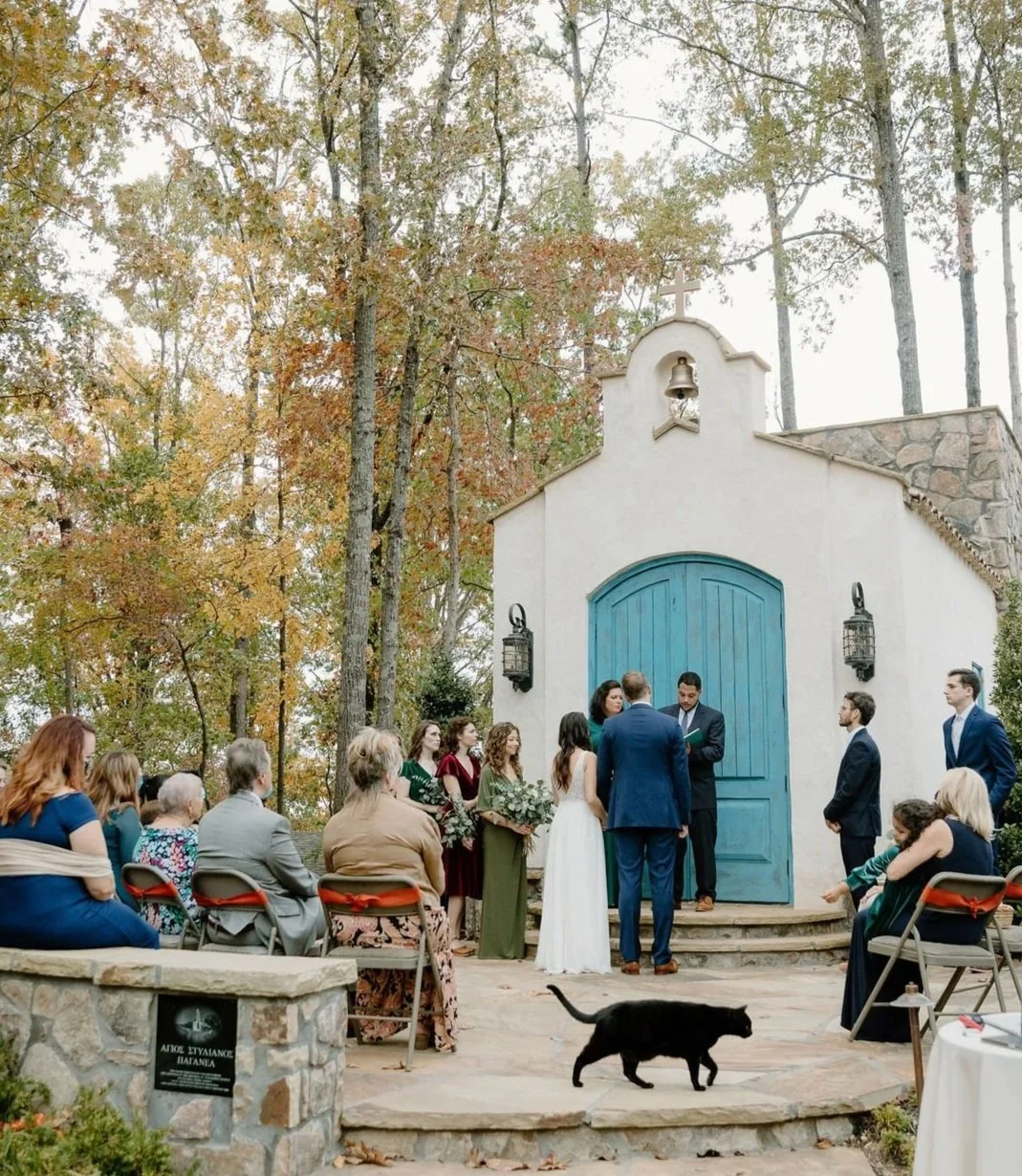 An outdoor wedding ceremony with the bride and groom exchanging vows in front of a small chapel surrounded by trees with fall foliage.